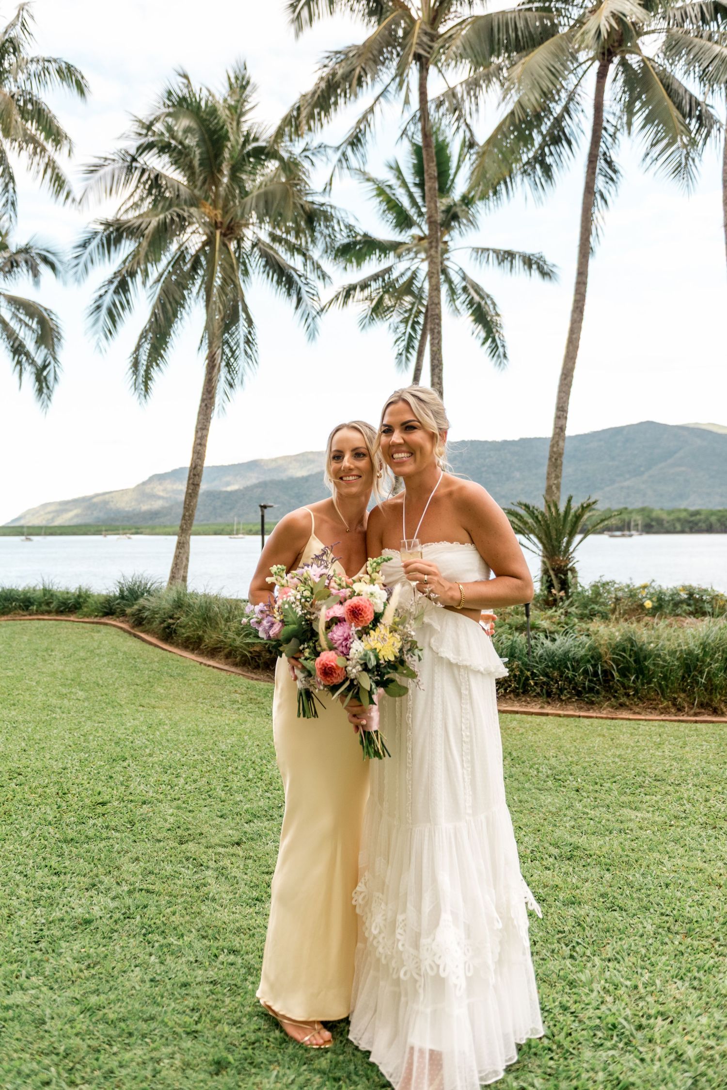 Bride in white lace gown and bridesmaid in cream dress smile holding bouquets near palm trees.