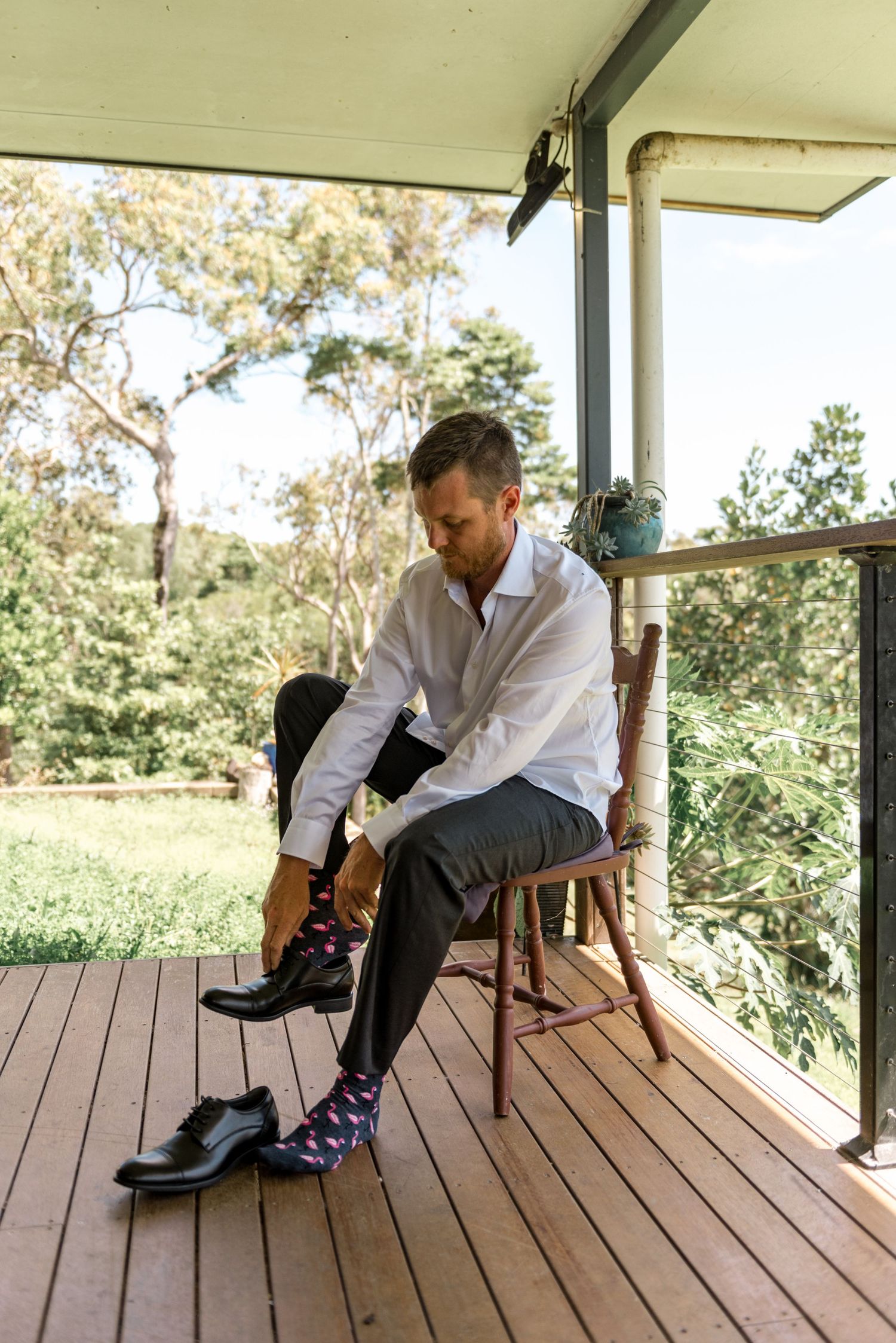 Groom sitting on a wooden chair on a porch putting on patterned socks and black dress shoes.