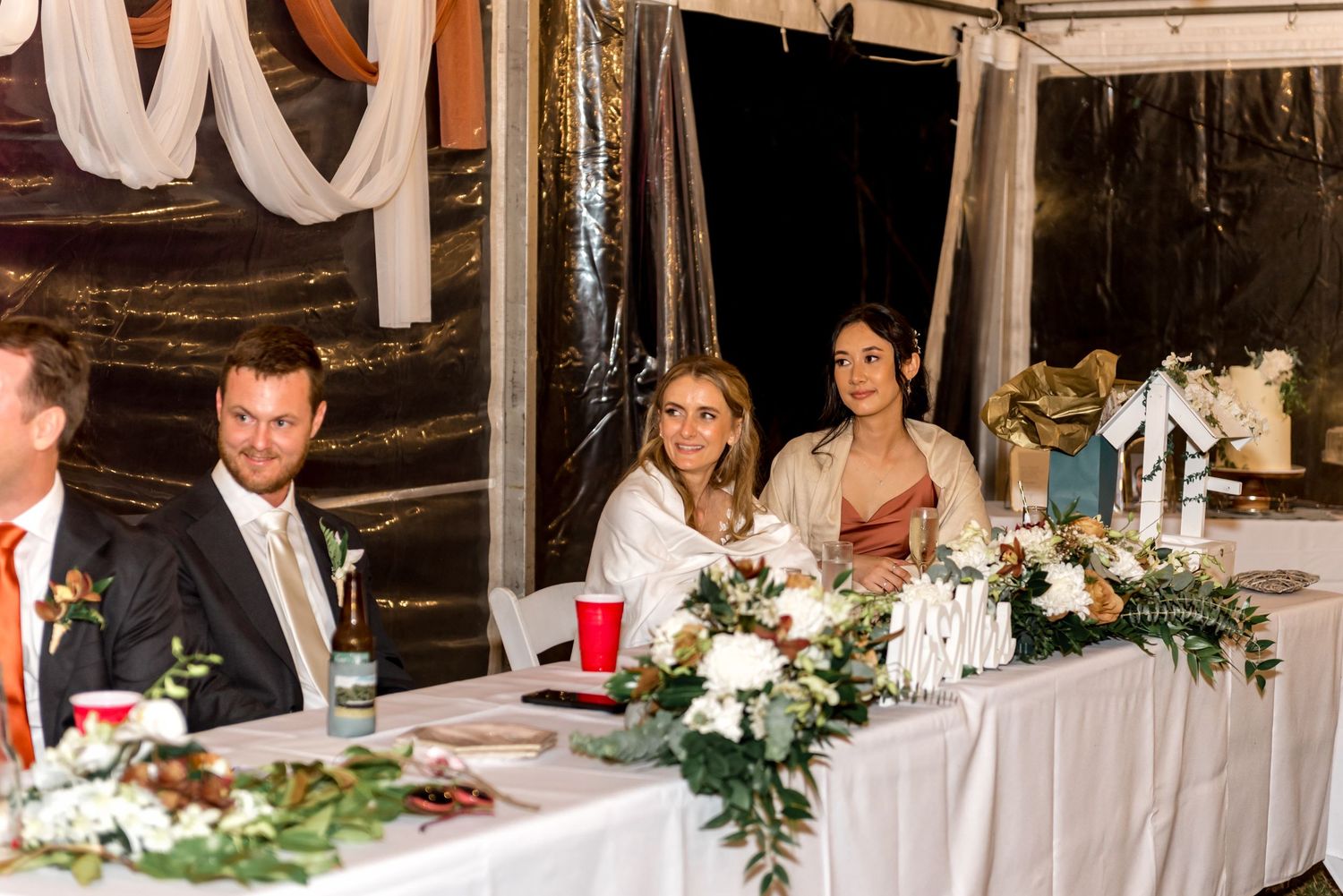 Wedding party seated at a decorated head table inside a tent with white floral arrangements and draped fabric.