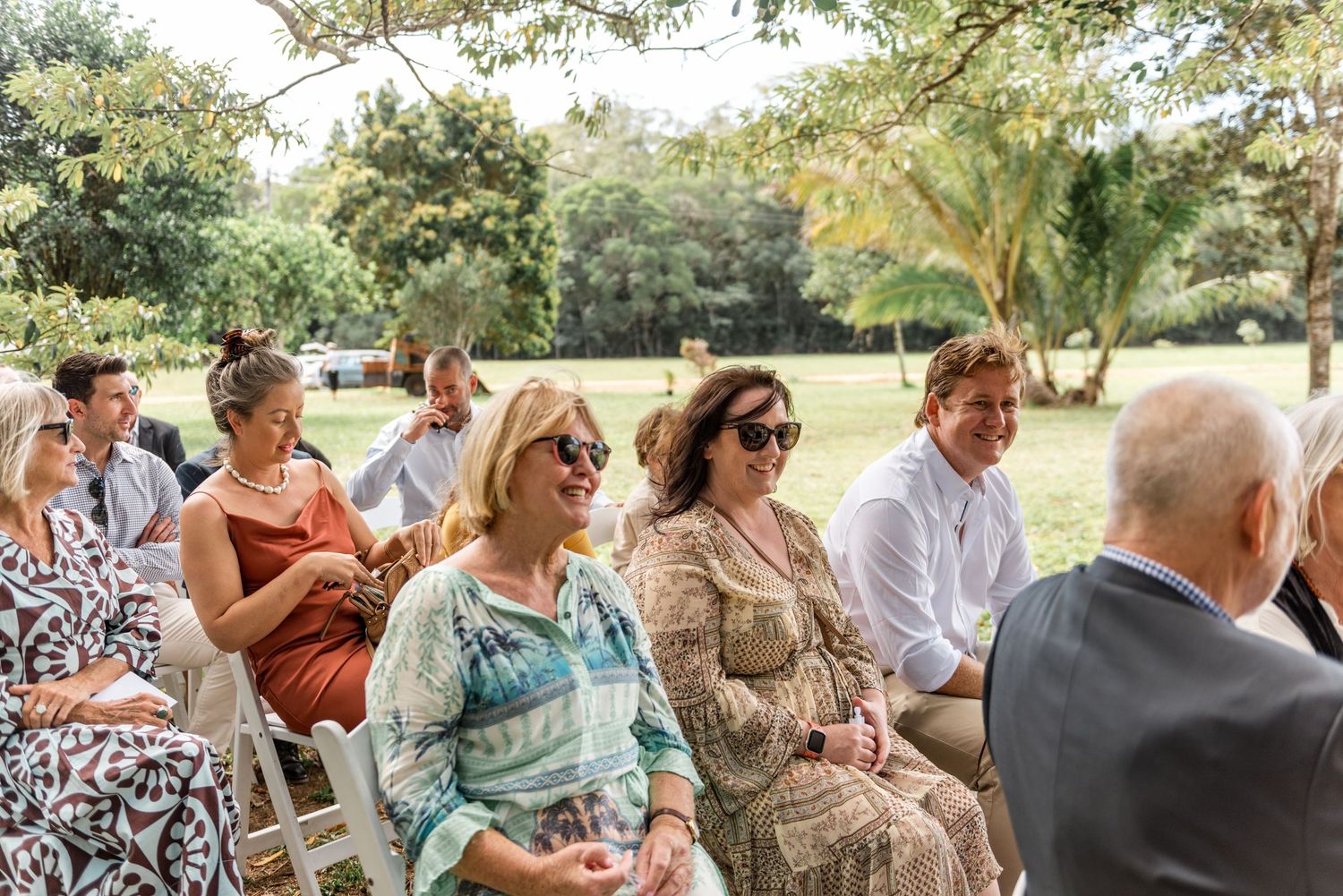 Smiling wedding guests seated outdoors under trees enjoying a sunny garden wedding ceremony.