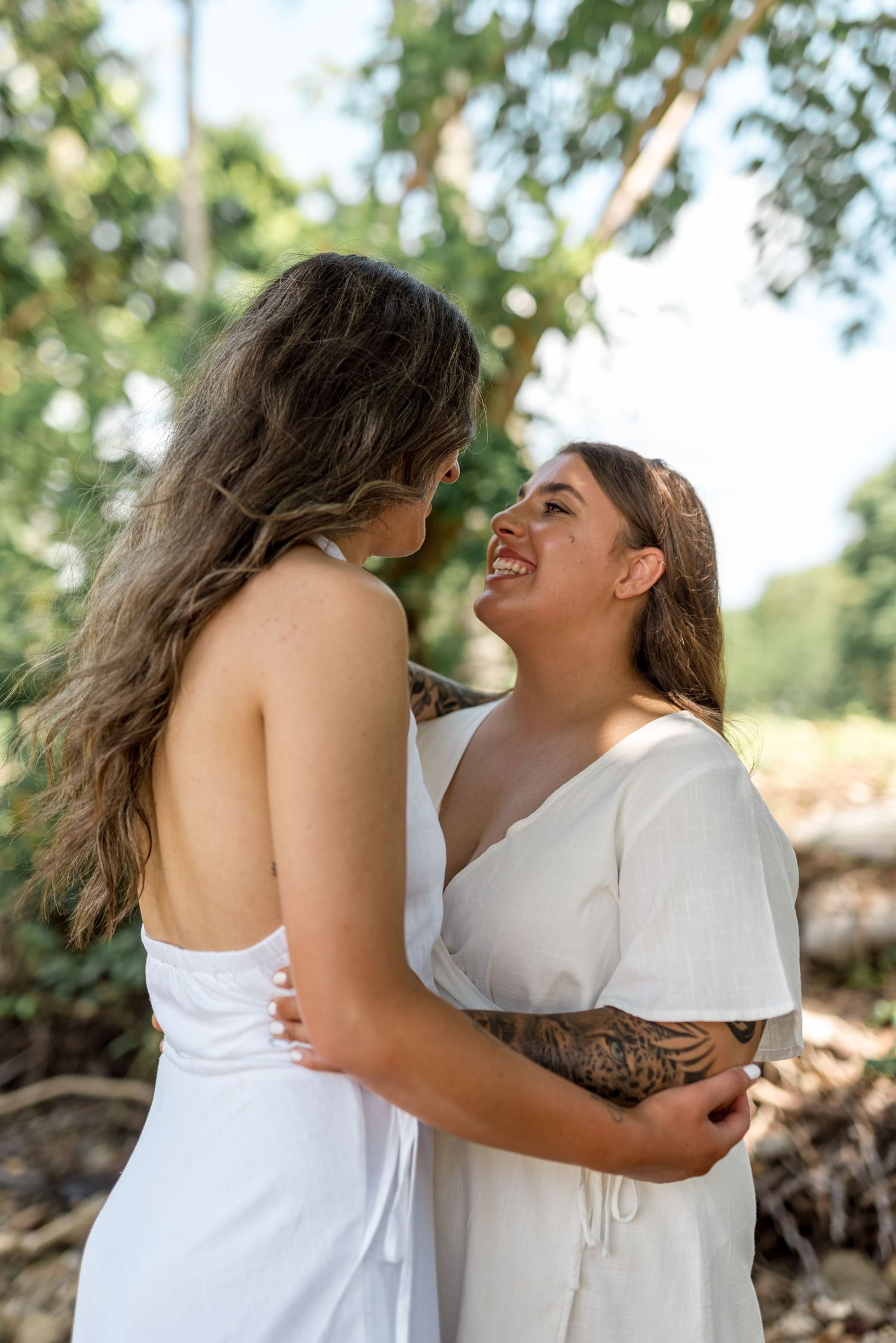 Two women in white dresses embrace and laugh together outdoors in a sun-dappled natural setting with green trees.