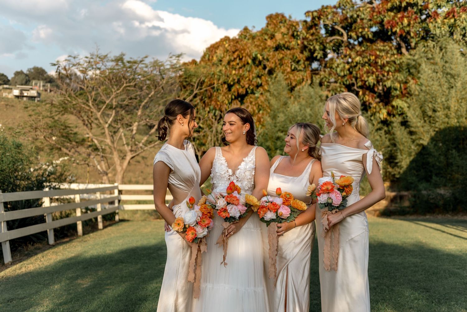 Bride and three bridesmaids in white dresses holding vibrant orange bouquets in a garden.