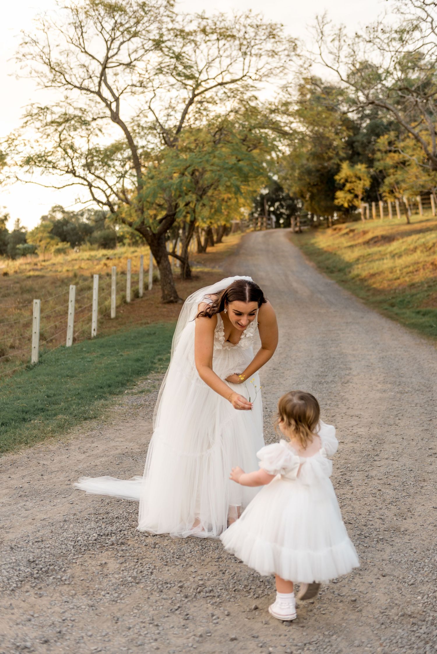 Bride in white tulle gown bends down to greet a toddler flower girl on a rustic country dirt road.