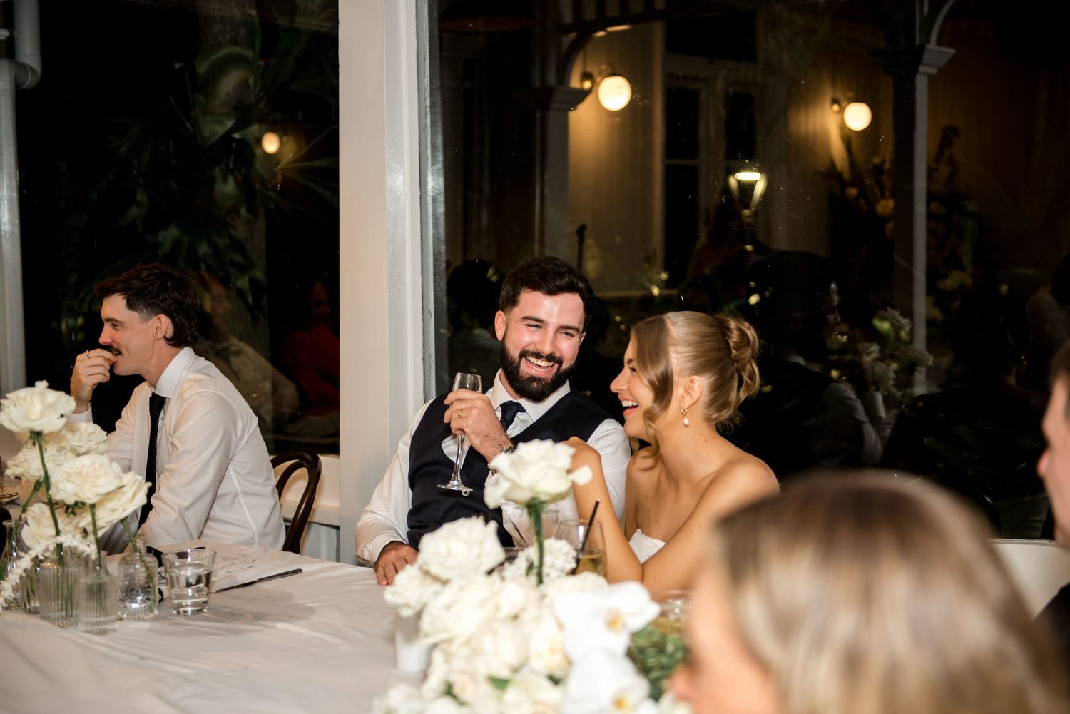 Smiling bride and groom seated at wedding reception table with white roses and candlelight.