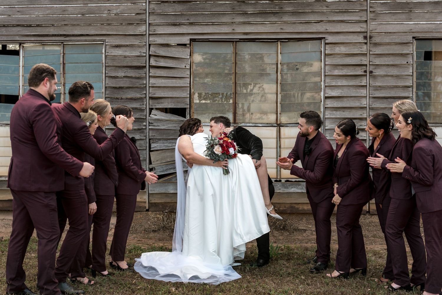 Bride and groom share a kiss surrounded by wedding party in matching burgundy suits outside a rustic wooden barn.