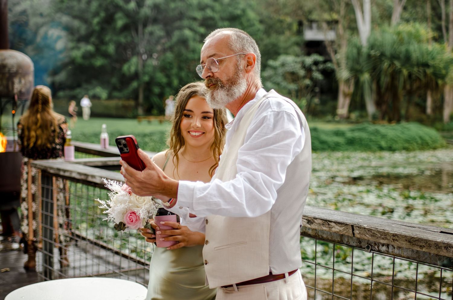 Smiling bride in sage green dress looks at phone with bearded man in white suit at outdoor wedding.