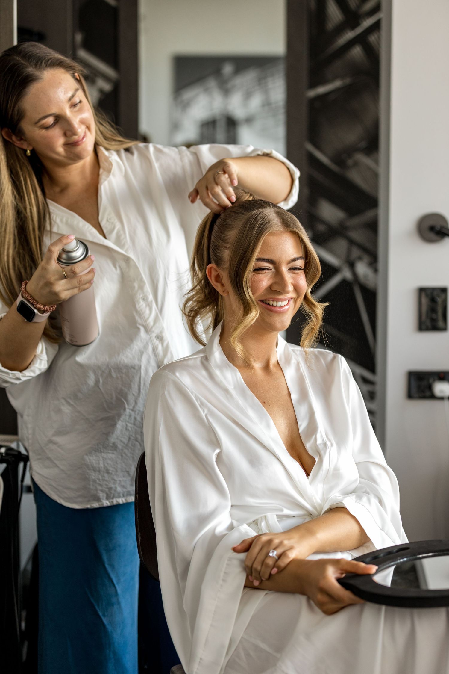 Smiling bride in white satin robe laughing as hairstylist applies hairspray during wedding preparations.
