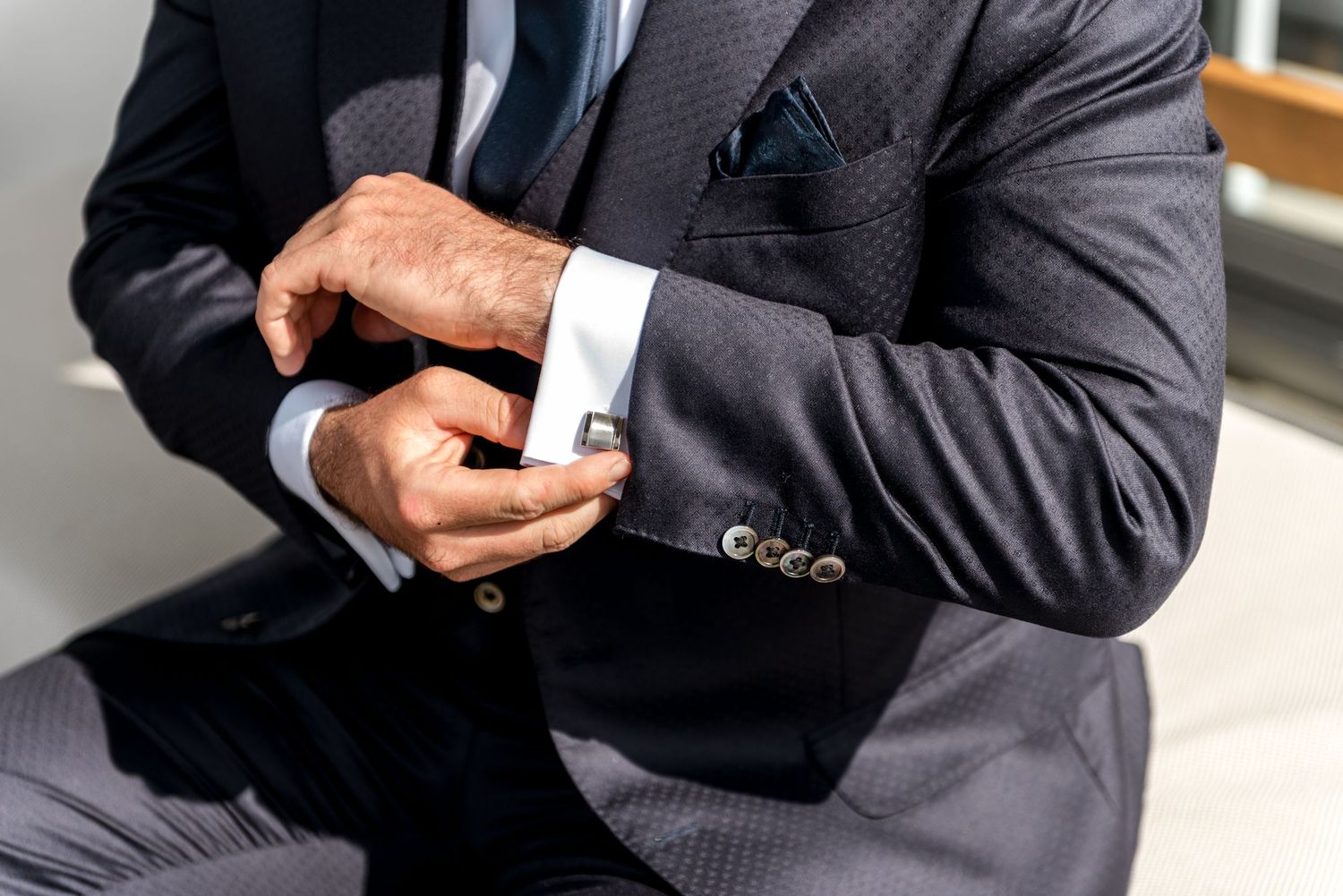 Close-up of a groom in a dark suit fastening silver cufflinks on his white dress shirt.