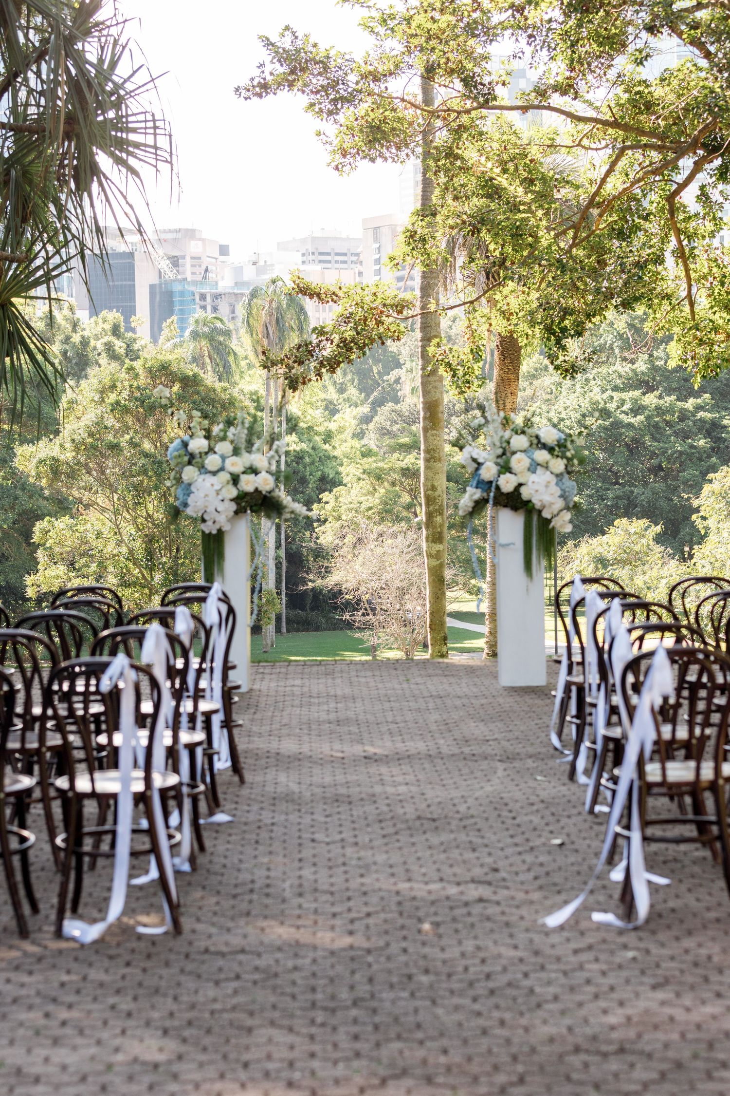 Outdoor wedding ceremony aisle with wooden chairs, white ribbon bows, and floral arrangements leading to a garden altar.