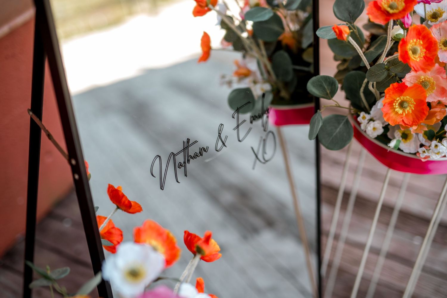 Close-up of a wedding mirror sign with Nathan and Emmy written in elegant script, surrounded by vibrant orange flowers.