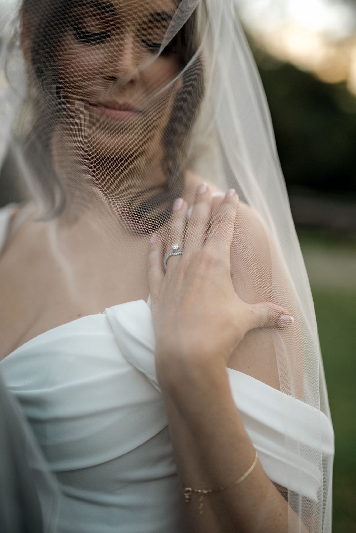 Bride in white strapless gown displaying diamond engagement ring and gold bracelet beneath flowing sheer veil.
