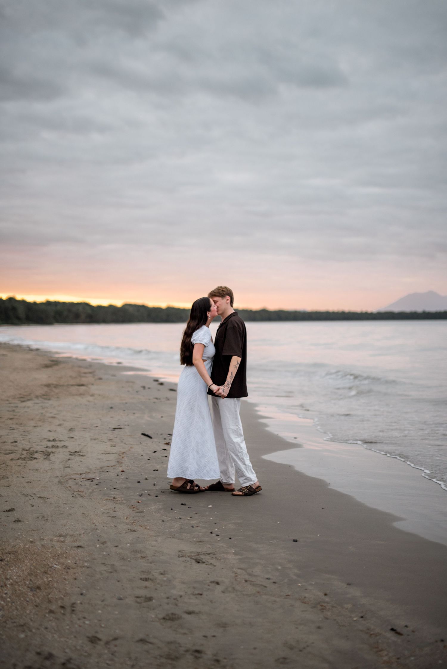 Couple shares romantic kiss on sandy beach at sunset with ocean backdrop.