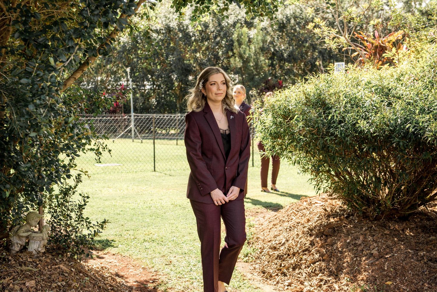 Elegant woman in burgundy suit walks through a sunny garden path at an outdoor wedding venue.