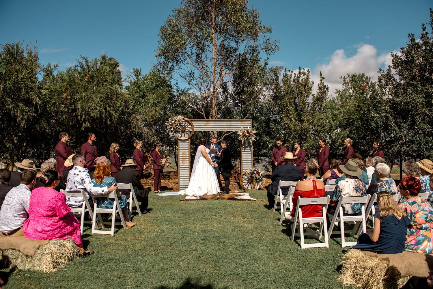 Wide shot of outdoor wedding ceremony on green lawn with guests seated on white chairs and hay bales.