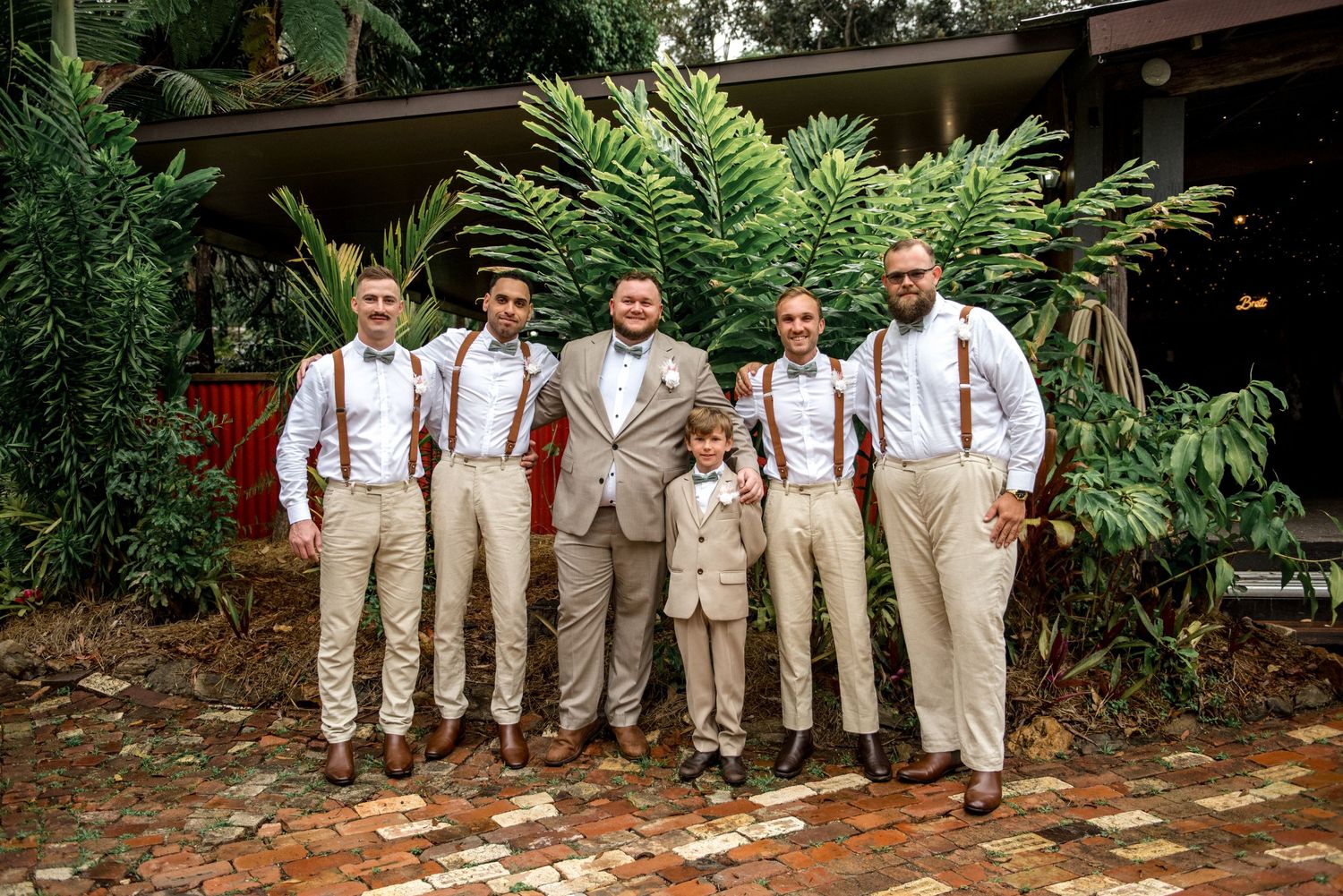 Groom and groomsmen in tan pants and brown suspenders posing outdoors at a wedding.