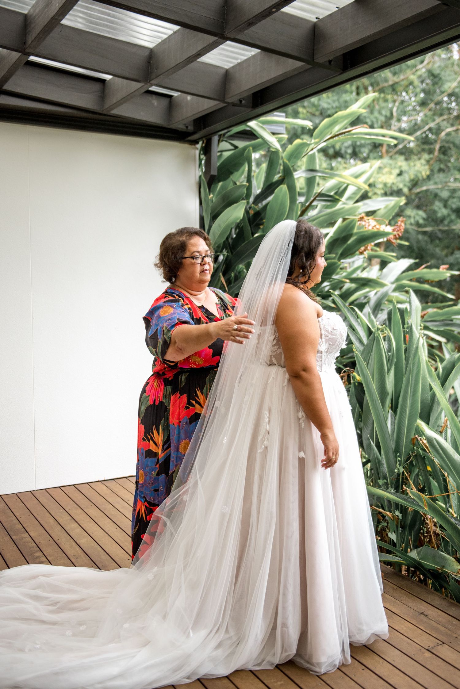 Mother adjusting the veil of bride in a flowing white wedding dress on a wooden deck outdoors.