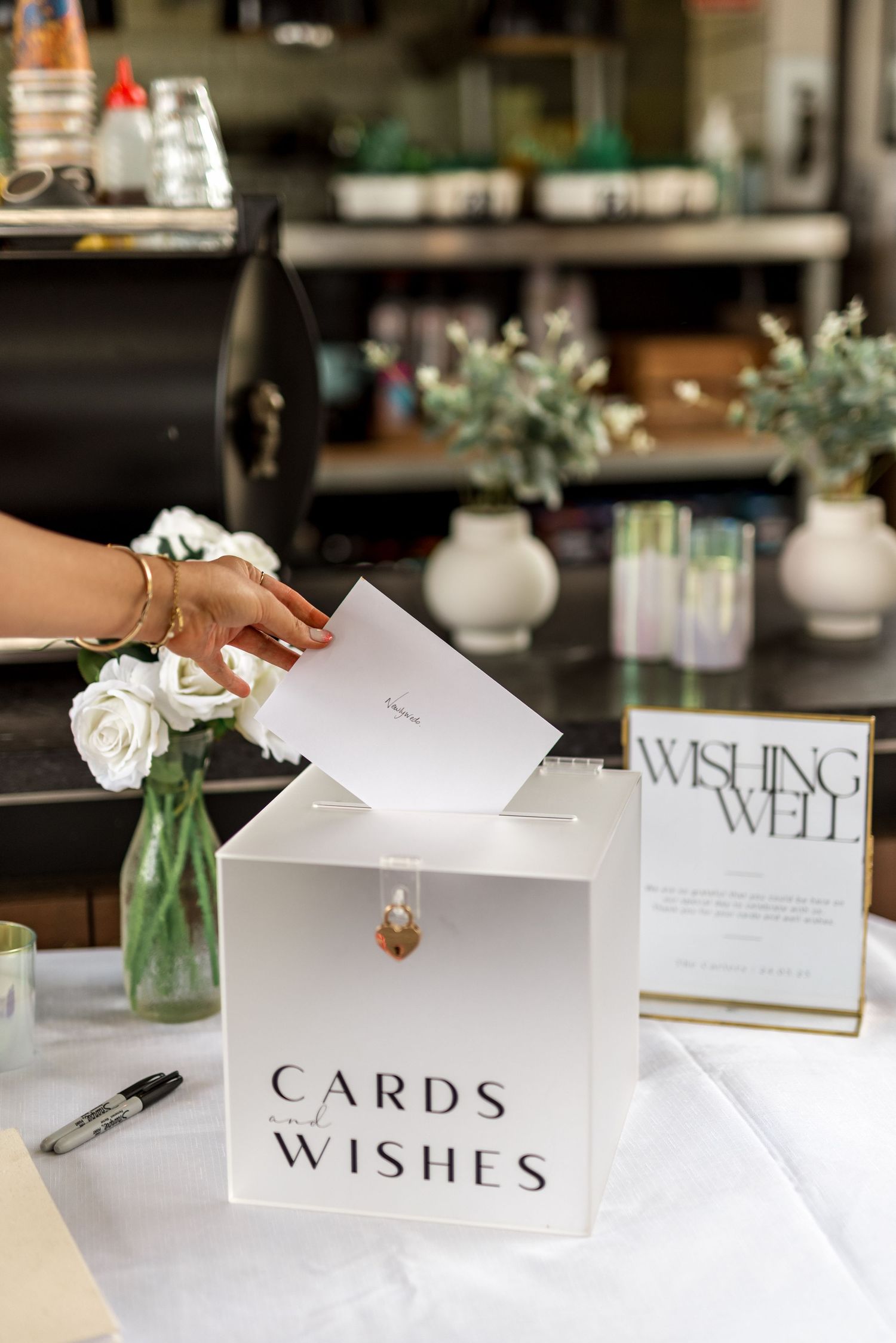 Hand placing a card into a white Cards & Wishes box at a wedding reception with a Wishing Well sign nearby.