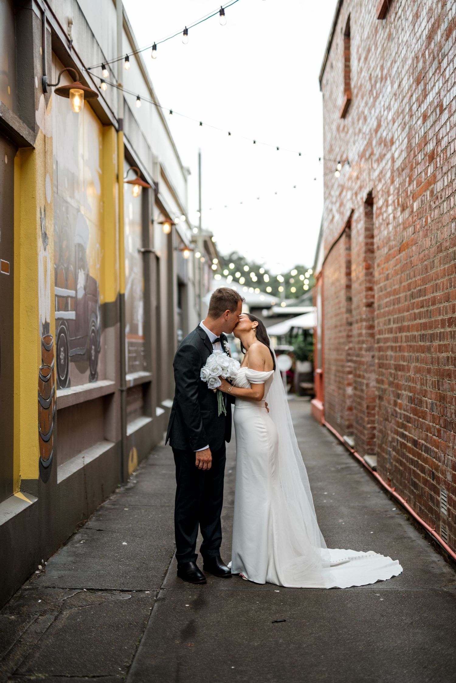 Bride and groom sharing a romantic kiss in a festoon-lit brick laneway.