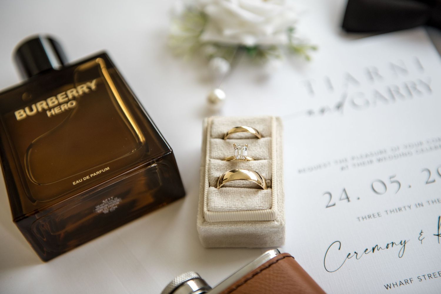 Close-up of gold wedding rings in a velvet box beside Burberry Hero cologne and a wedding invitation for Tiarni and Garry.