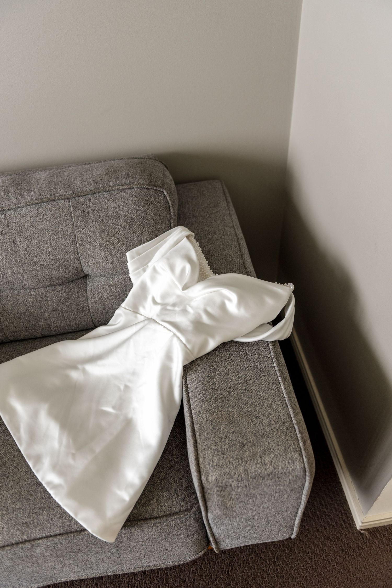 White satin strapless wedding gown casually draped over grey upholstered sofa in bridal preparation room.