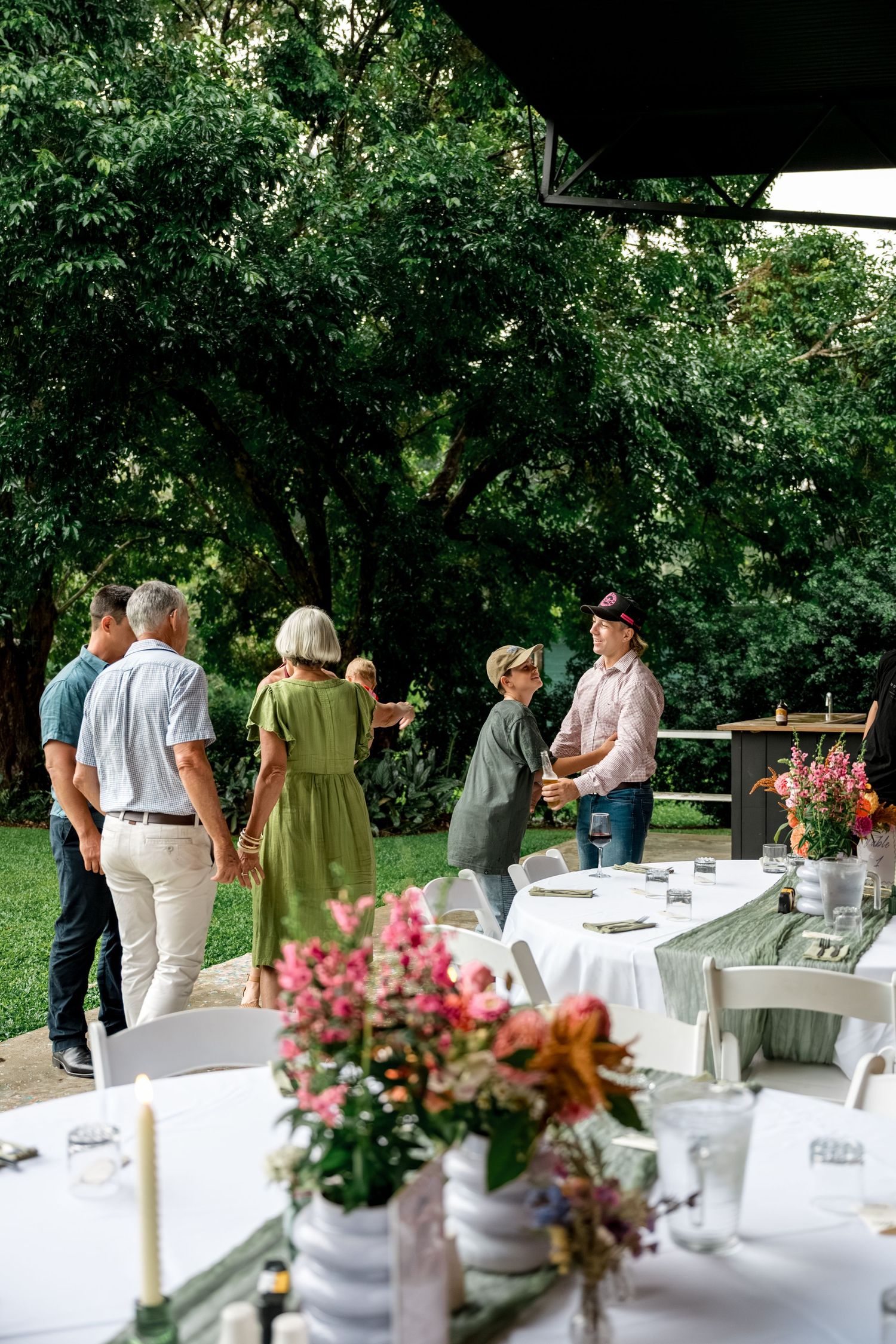 Guests mingle at an outdoor garden wedding reception with white chairs and floral centerpieces.
