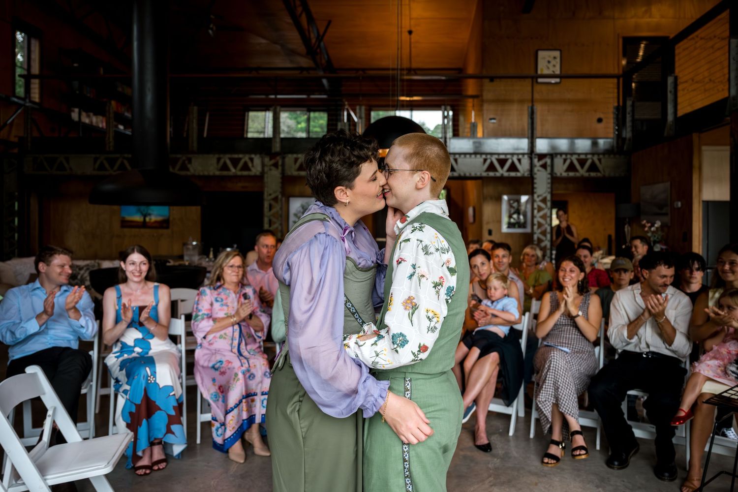 Two brides share first kiss at indoor wedding ceremony as guests applaud in background.