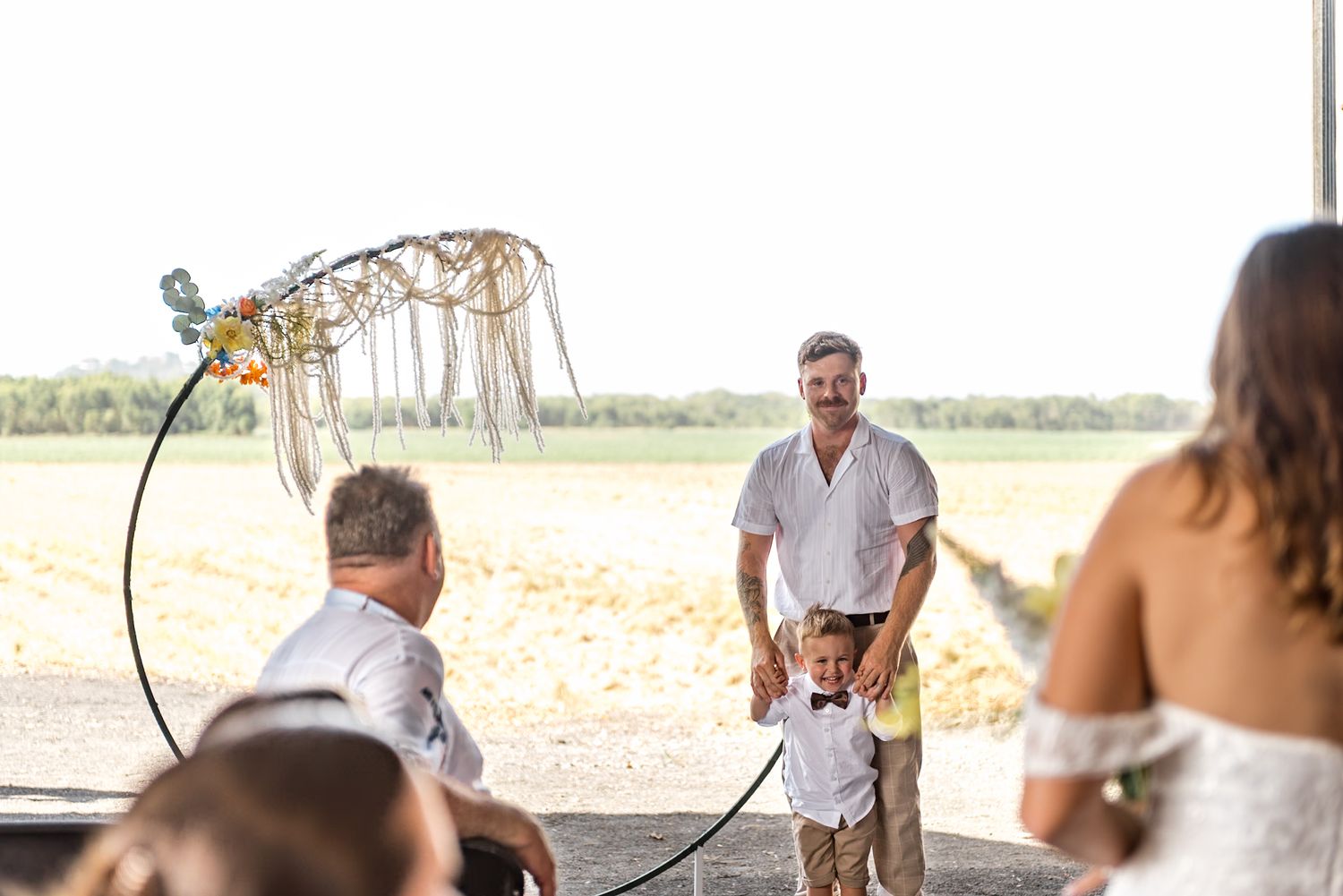 Groom smiles at young child during rustic barn wedding ceremony beneath a circular floral arch.