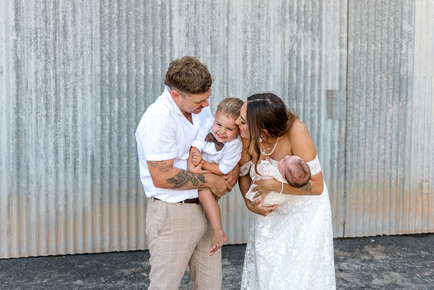 Newlywed couple in wedding attire kissing toddler son while bride holds sleeping newborn baby.