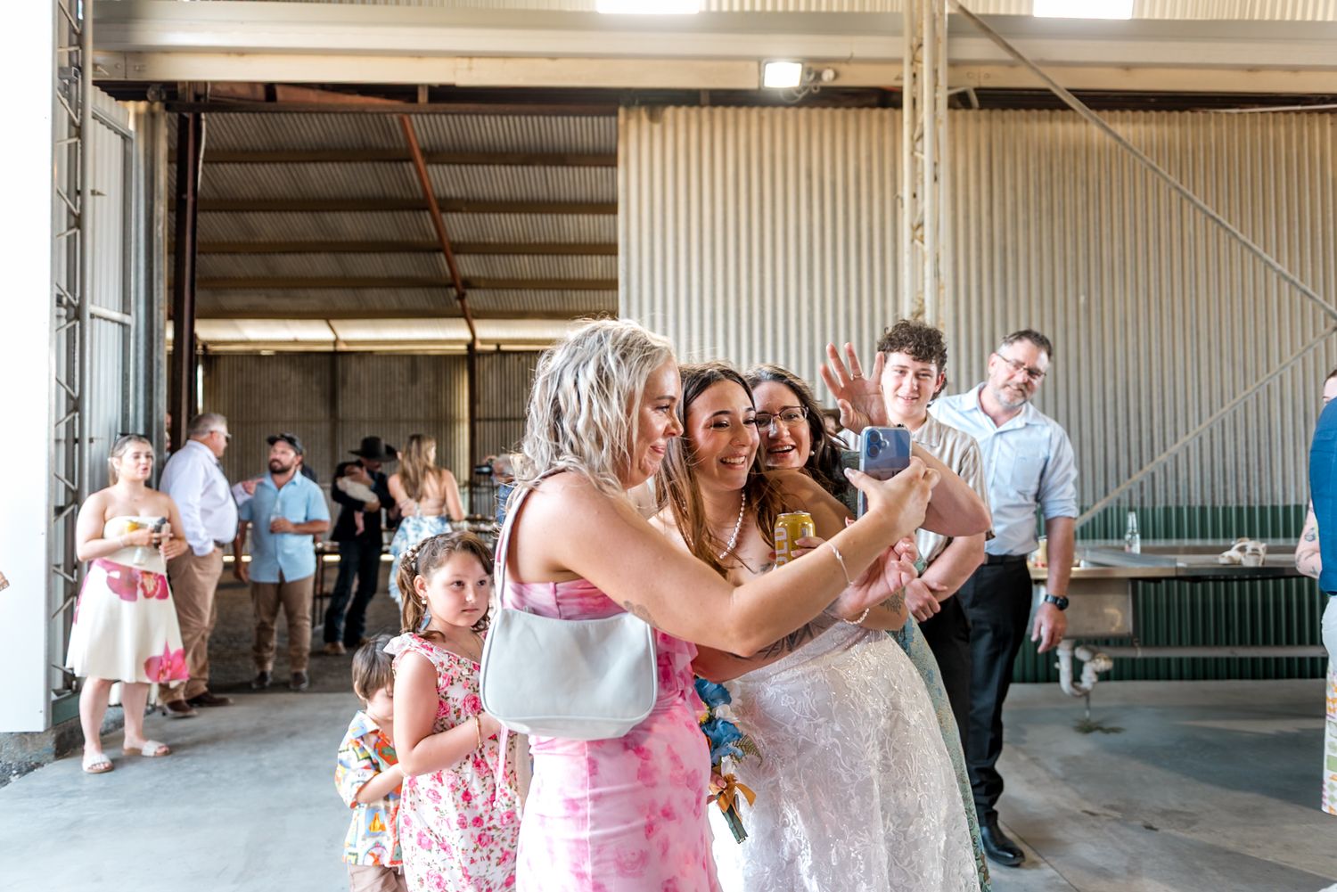 Bride in white lace gown embraces a guest in pink while children and wedding guests look on.
