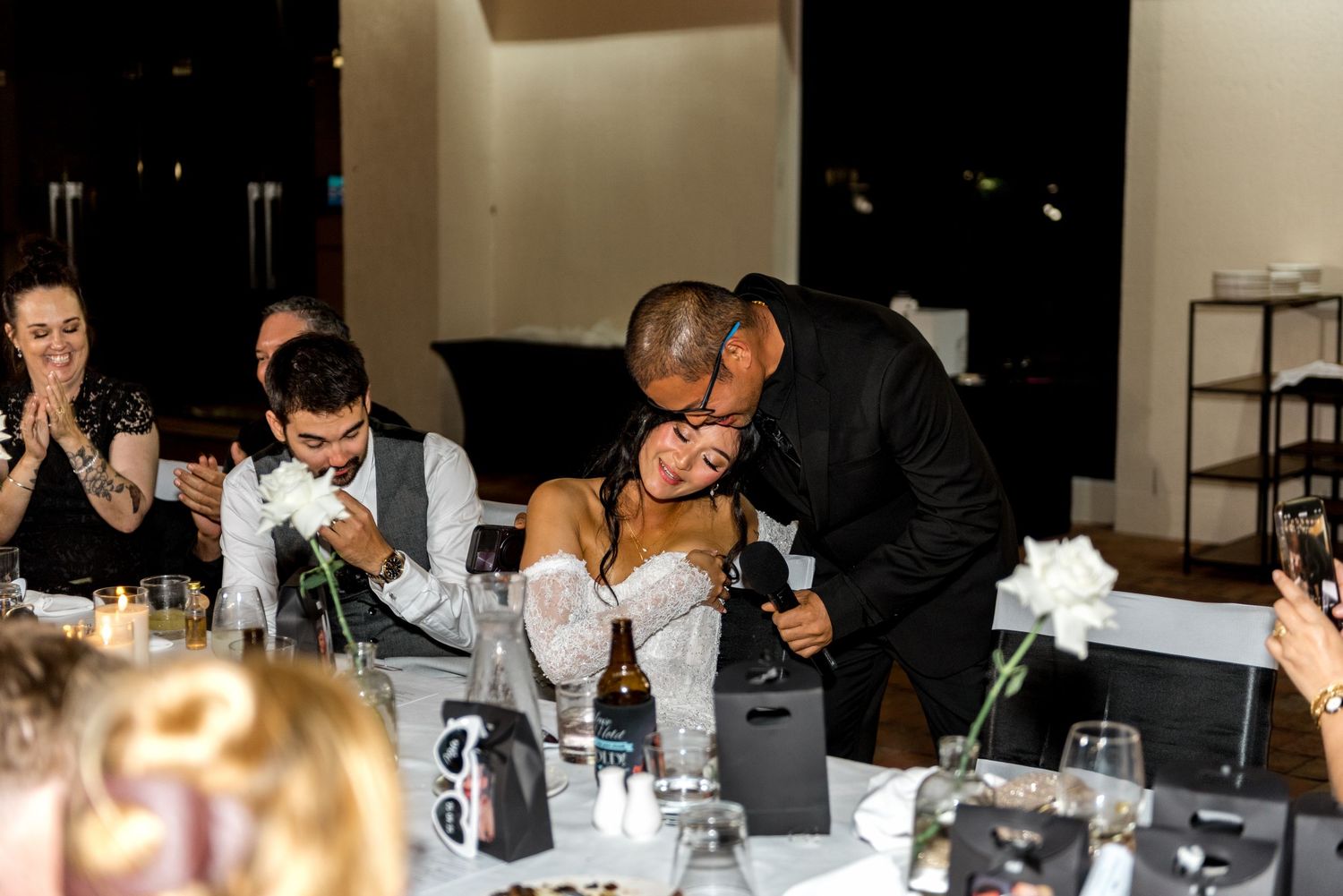 Bride receives a warm hug from a guest while seated at the head table during the indoor wedding reception.