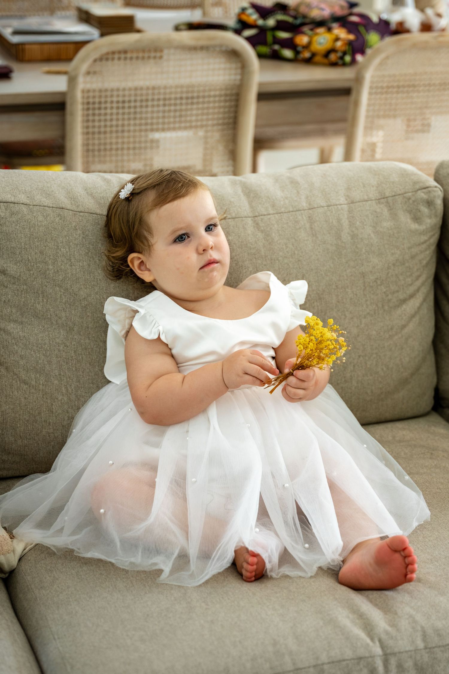 A baby girl in a white tulle dress holds yellow flowers while sitting on a beige sofa.