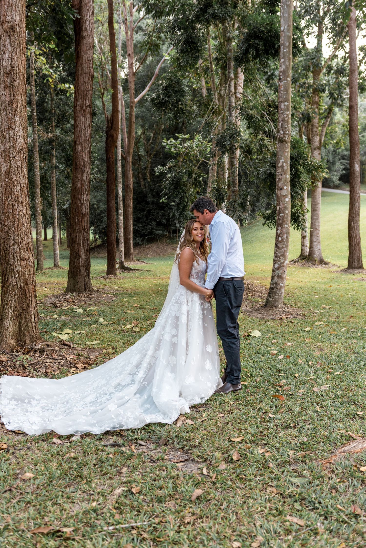 Bride and groom share a tender moment among tall trees, her lace gown train spread across the grass.