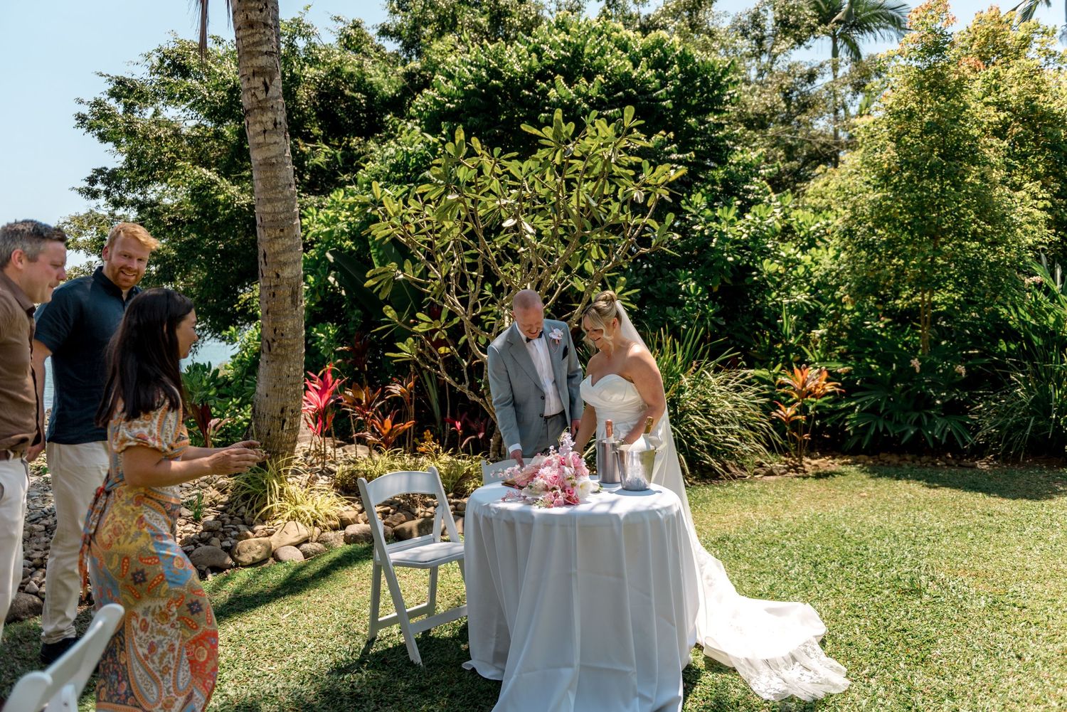 Newlyweds cutting wedding cake in lush tropical garden setting.