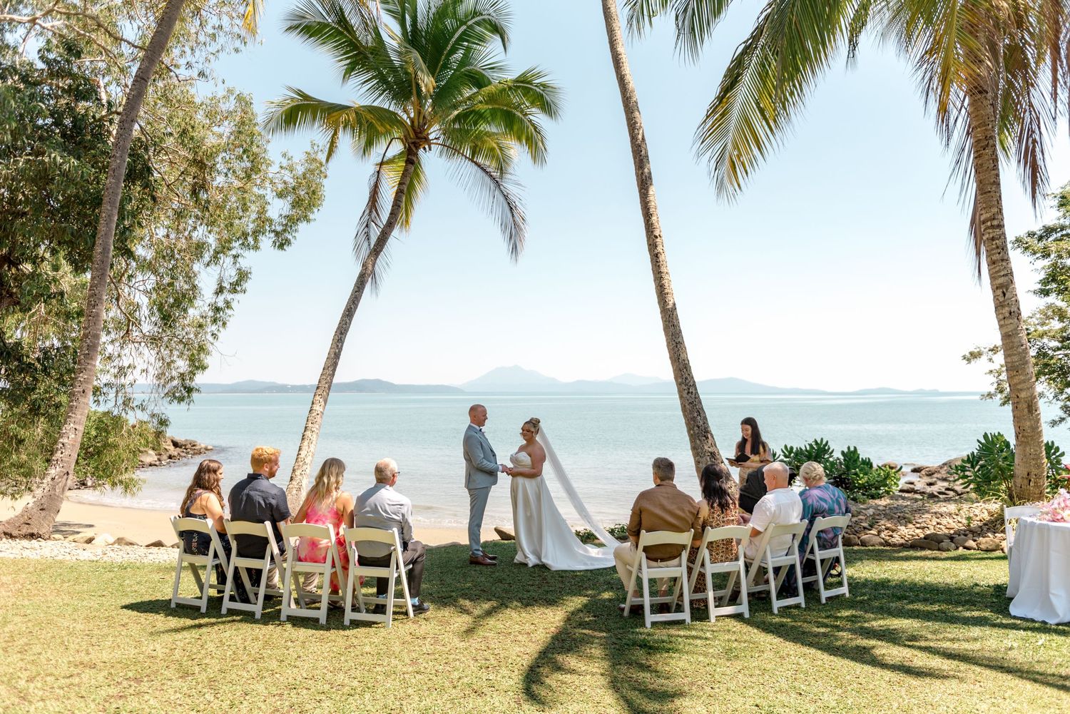 Intimate beach wedding ceremony under palm trees with small guest gathering and ocean backdrop.