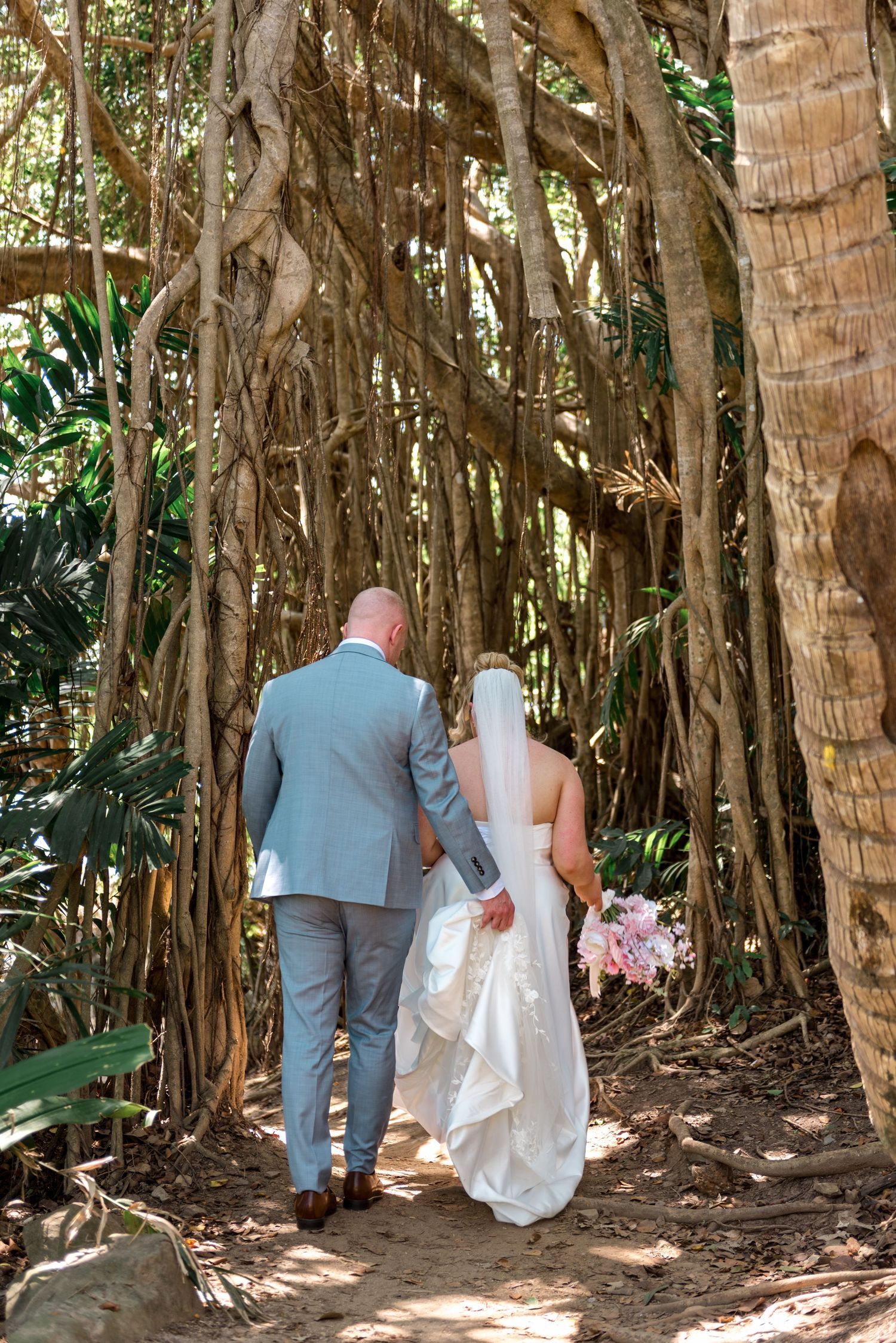 Bride and groom walk hand-in-hand through a lush tropical banyan tree forest on their wedding day.