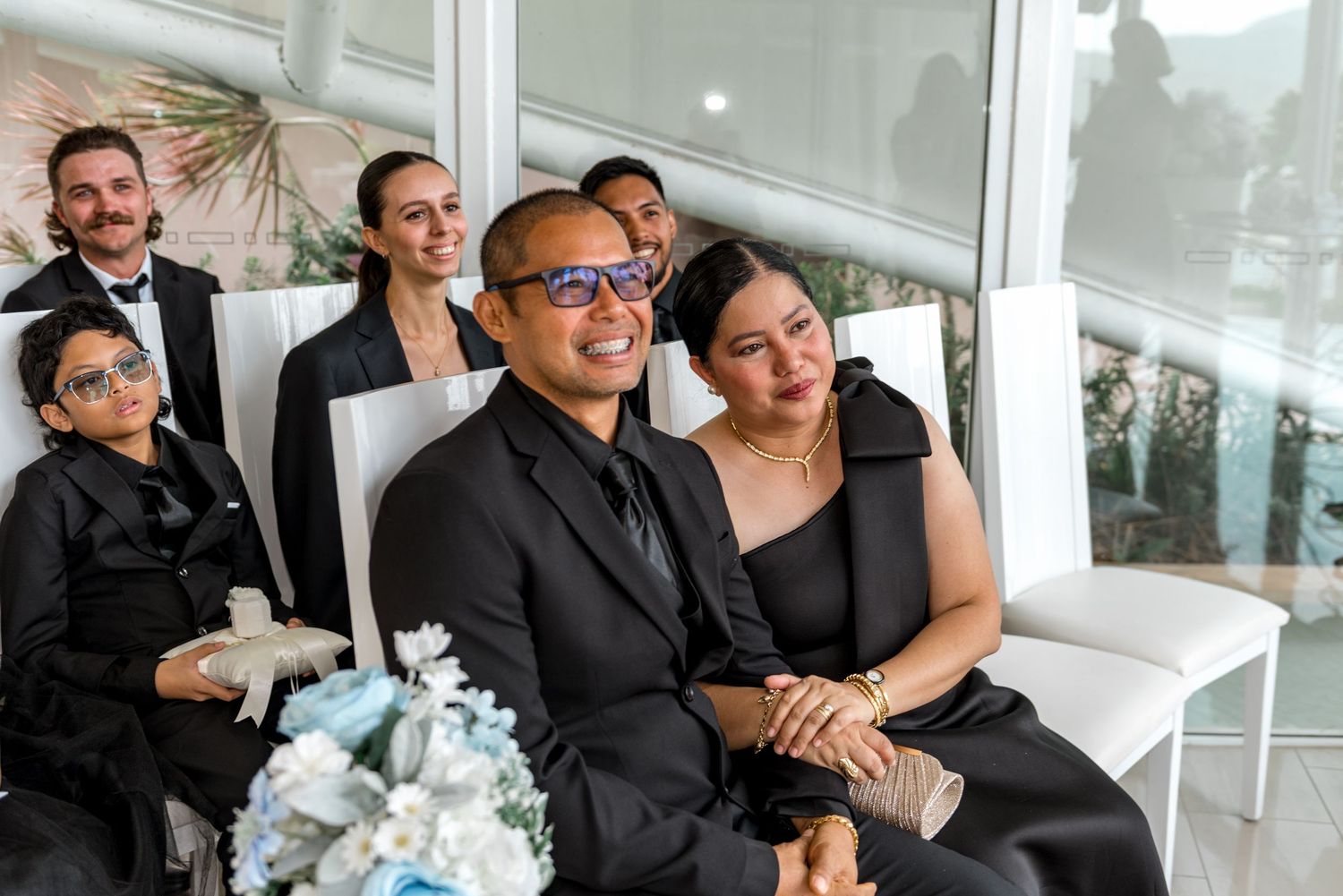 Smiling wedding guests in black attire seated together during ceremony, woman leaning on man's shoulder.