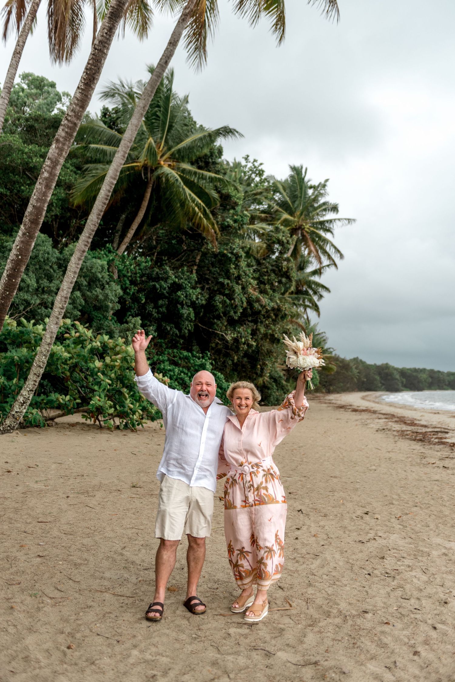 Joyful mature couple celebrating on a tropical beach with palm trees, raising their arms in excitement.