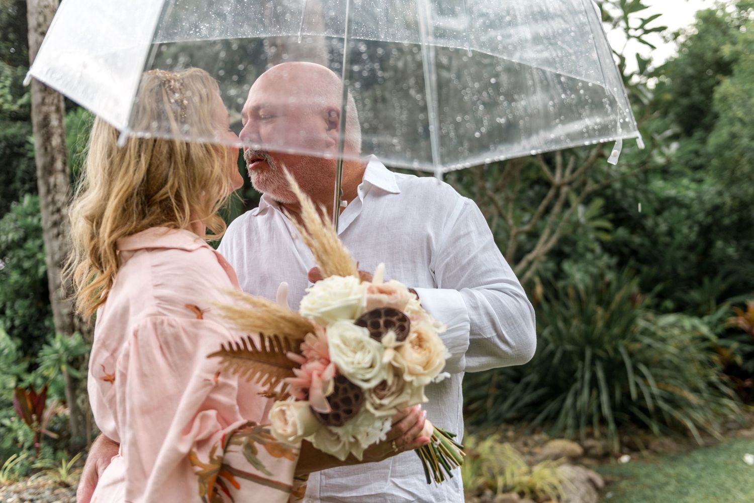 Romantic mature couple sharing an intimate moment under a clear umbrella with a bridal bouquet.