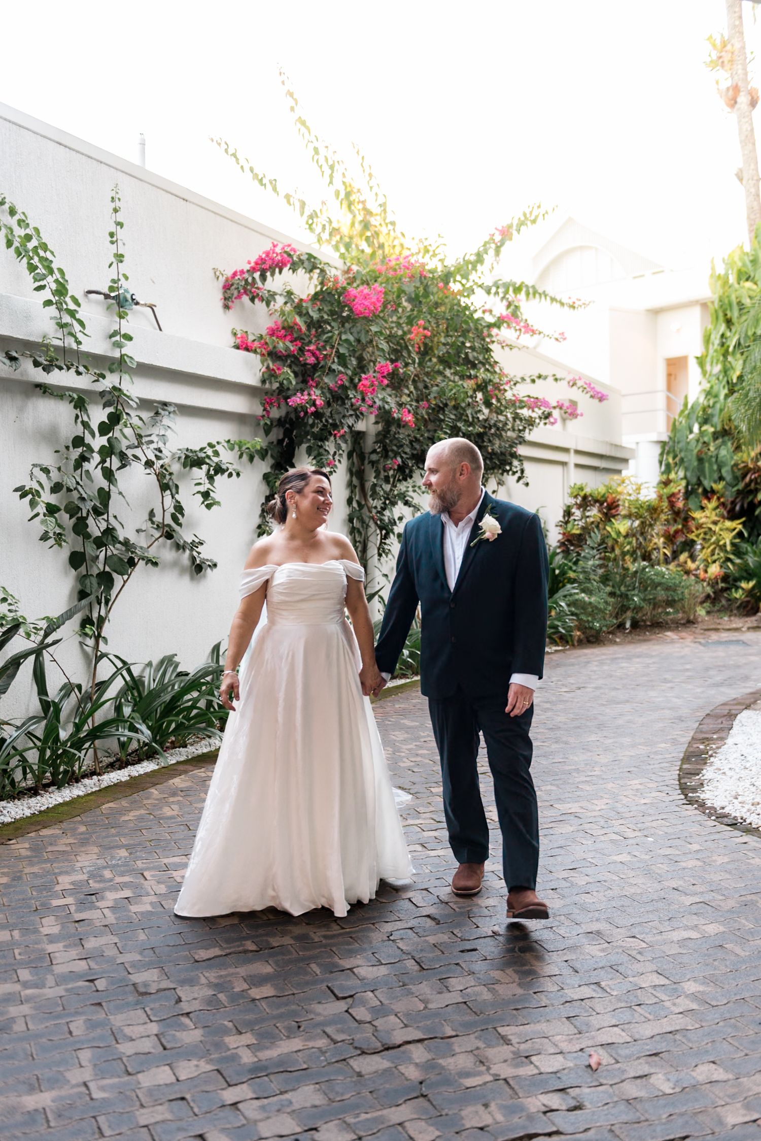 Bride and groom walking hand in hand on brick path with pink bougainvillea backdrop.