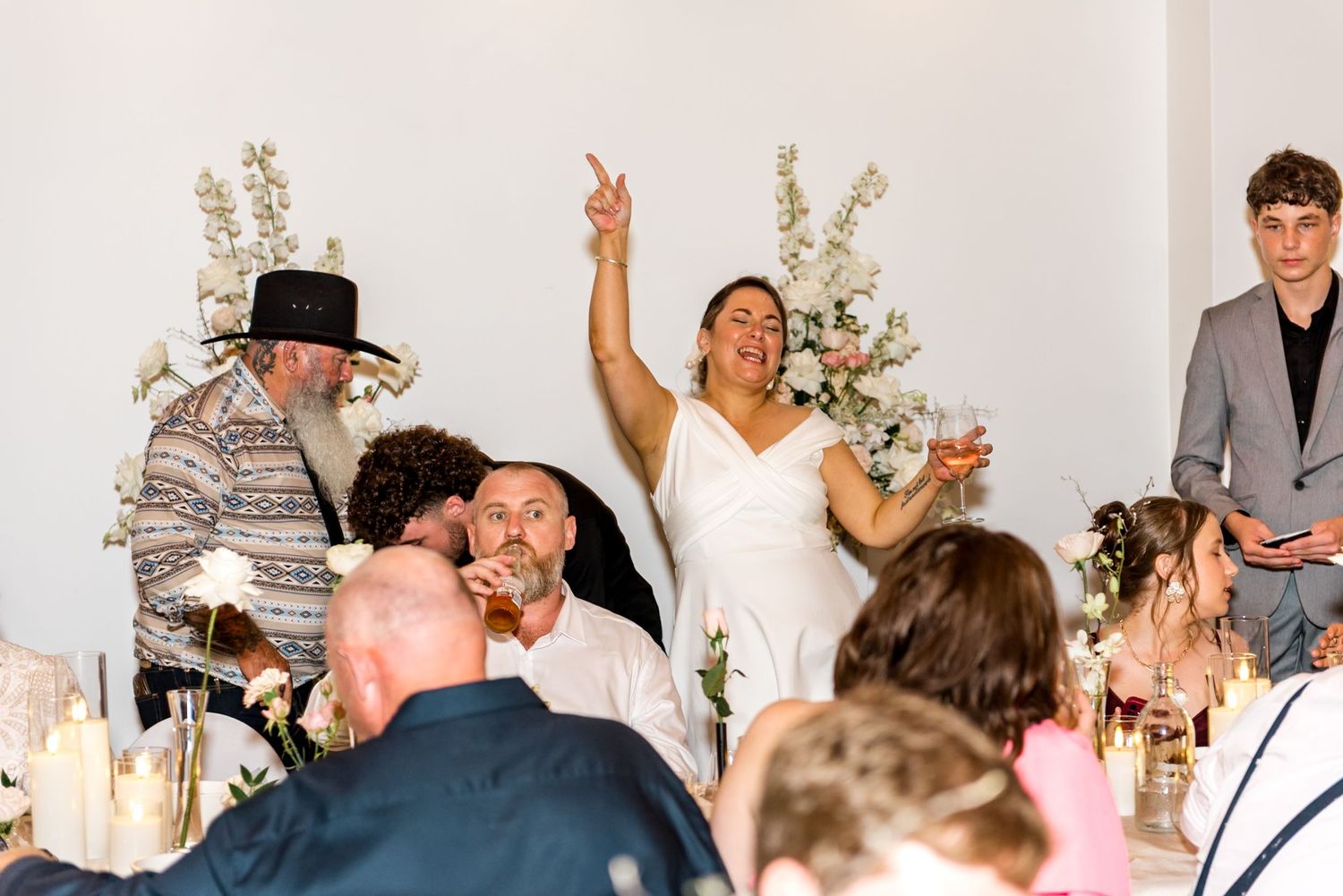 Bride in white dress celebrating joyfully with raised arm at wedding reception dinner.