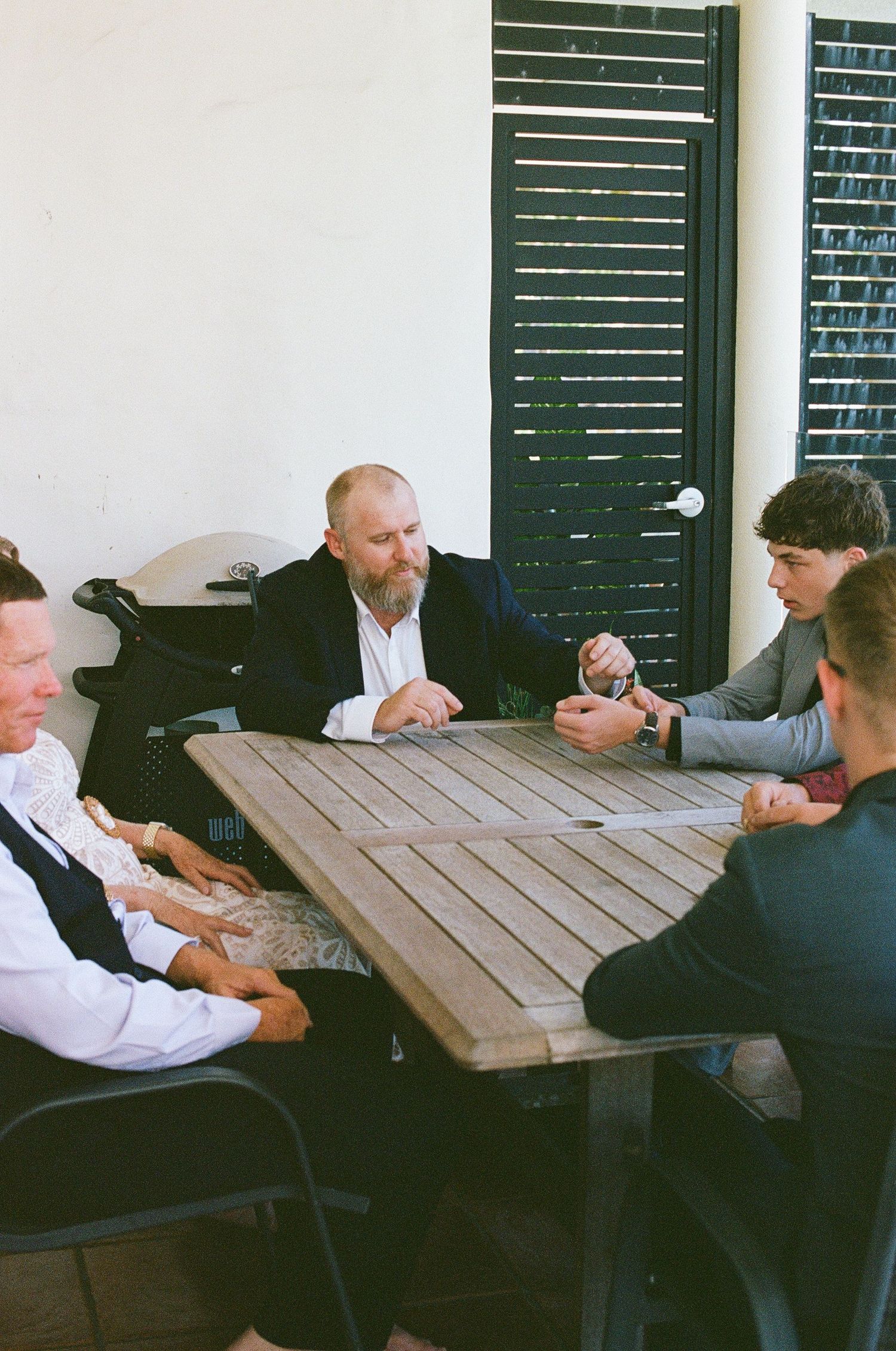 Group of men in suits sitting around wooden outdoor table in casual wedding conversation.