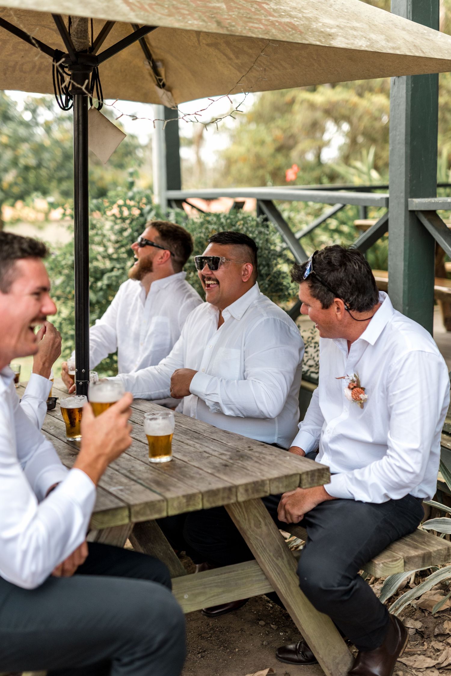 Groomsmen in white shirts relaxing with beers at an outdoor picnic table before the wedding ceremony.