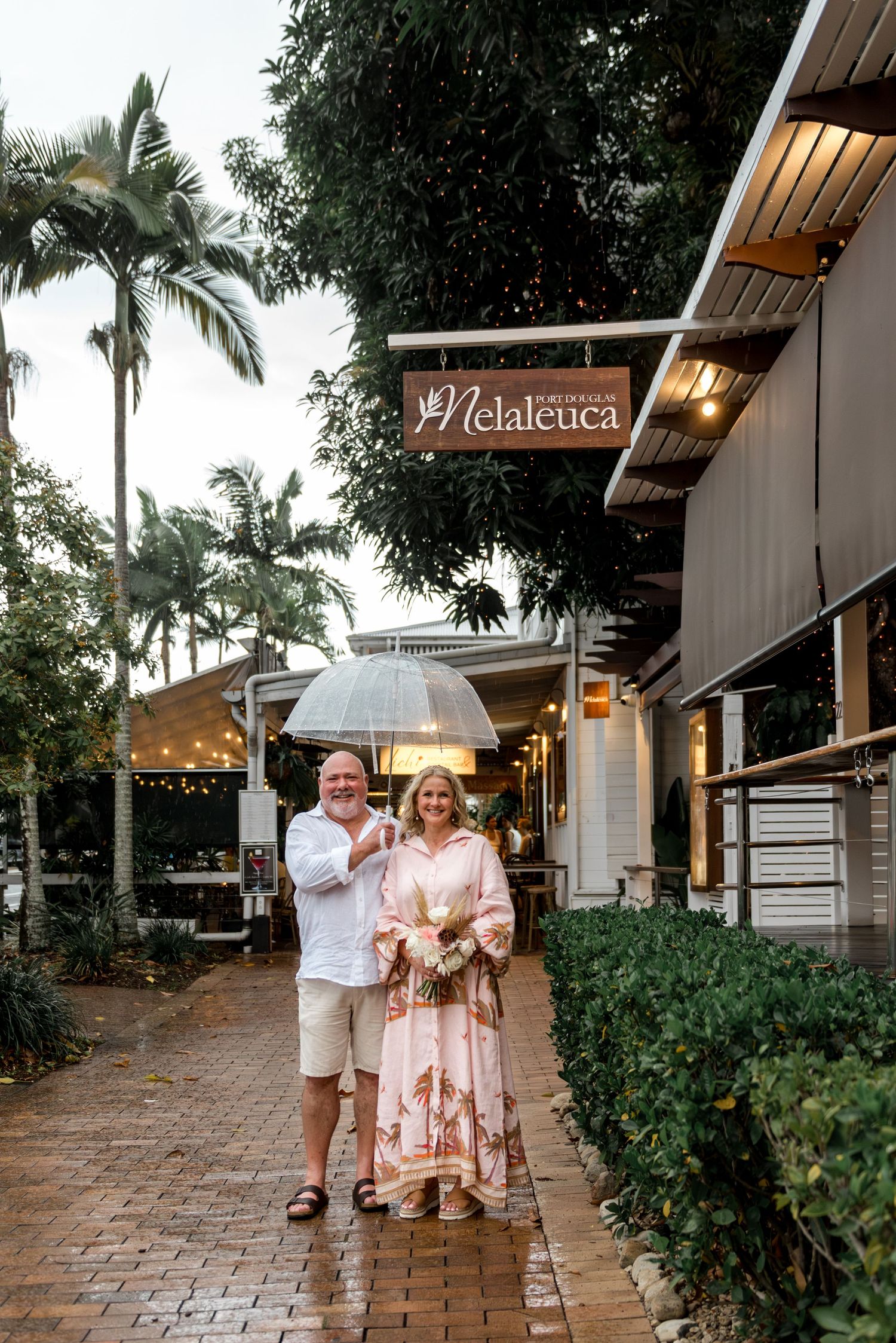 Couple smiling under clear umbrella outside Melaleuca restaurant on a rainy day.