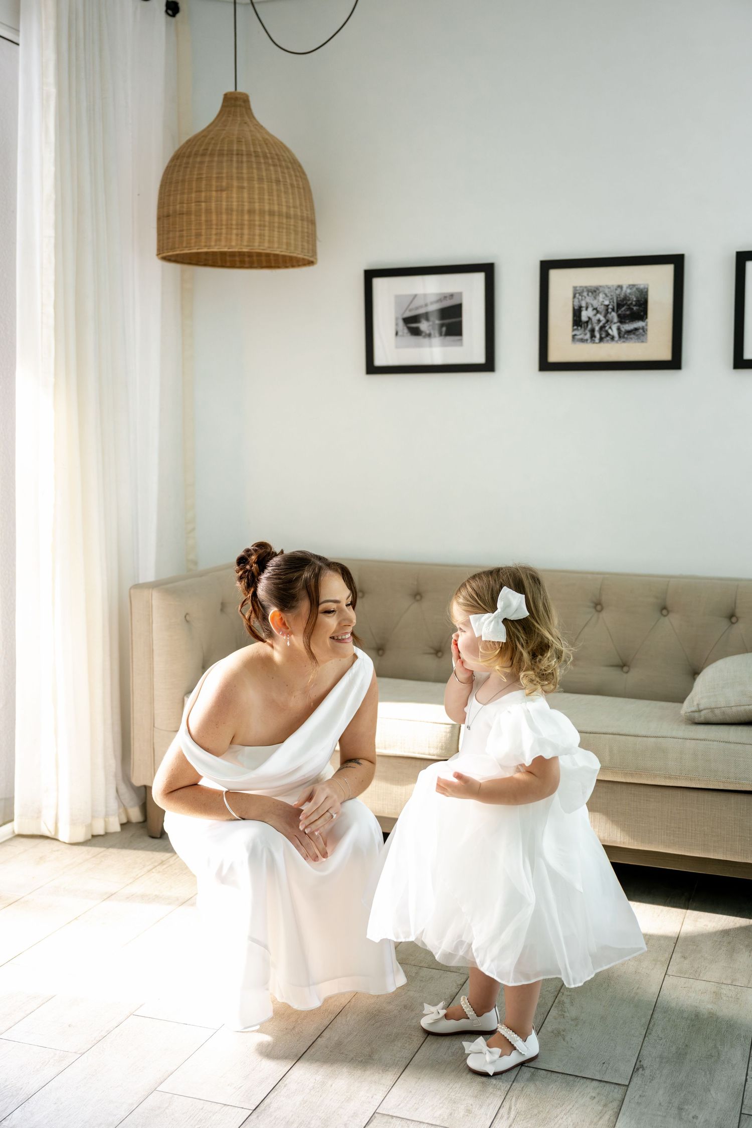 Smiling bride in white wedding dress interacting with adorable flower girl in white dress in sunlit interior at Sheraton Grand Mirage Resor.