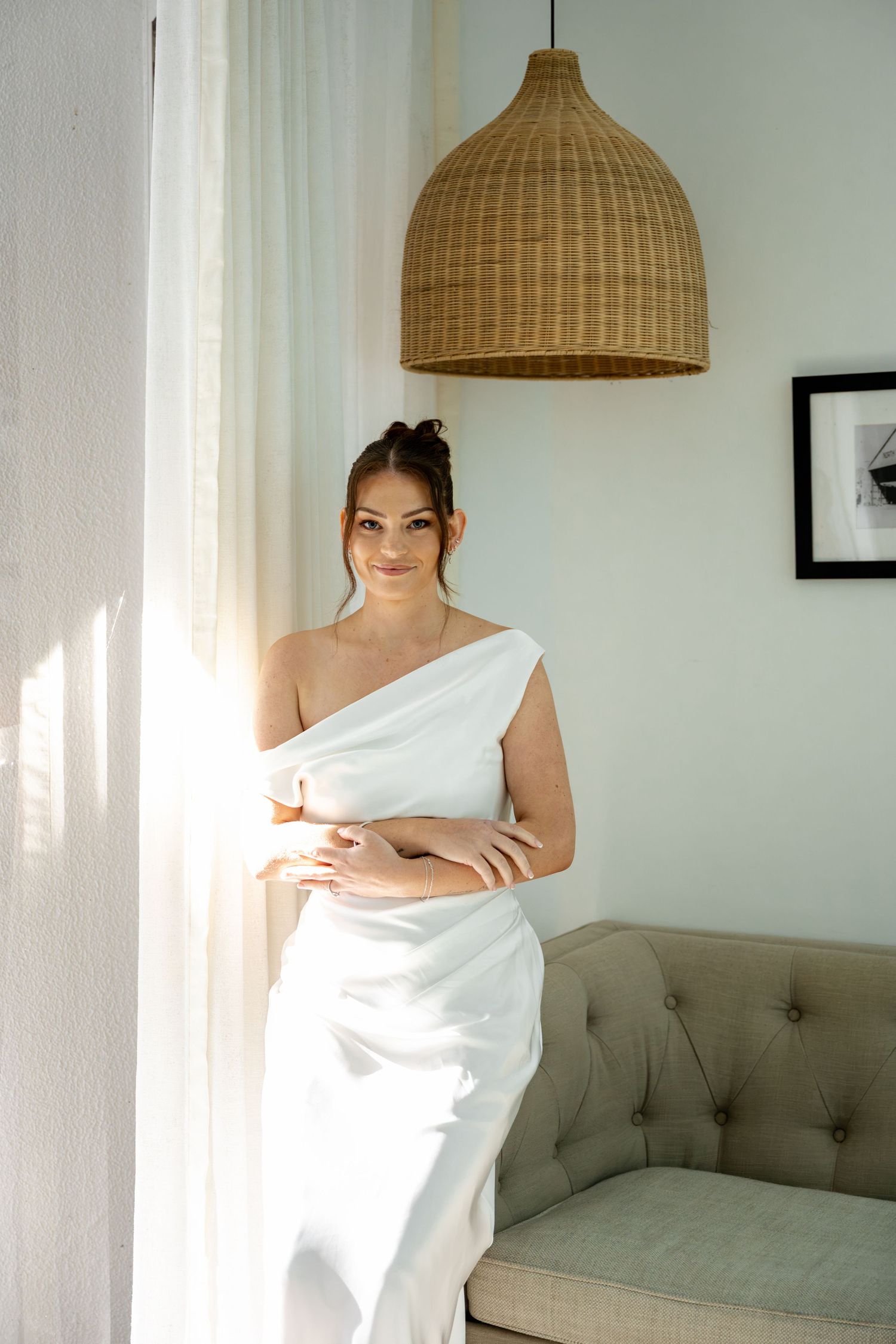 Smiling bride in white one-shoulder satin gown leans on sofa by sunlit window with rattan pendant light.