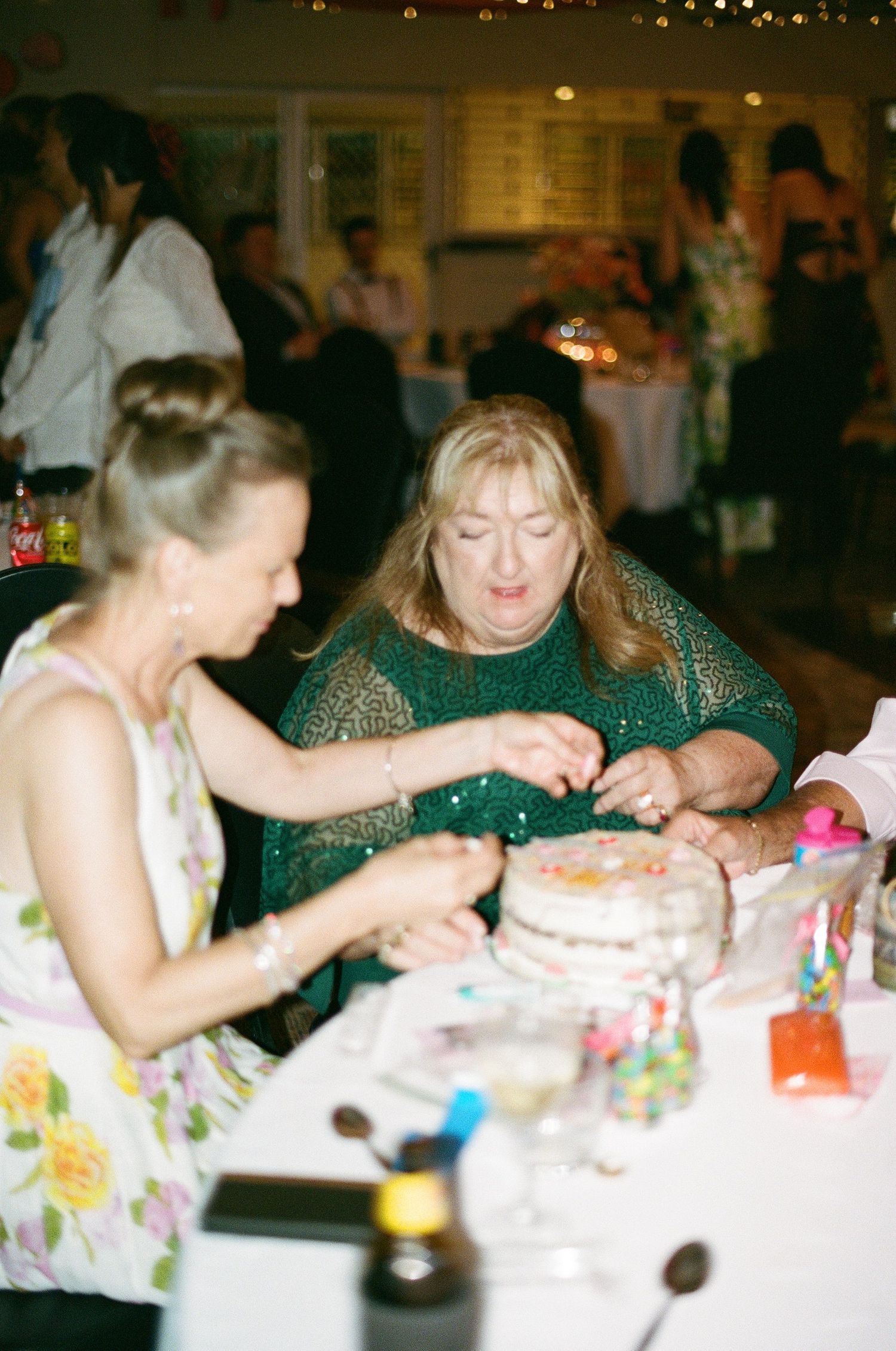 Two women decorating a layered wedding cake with colorful candies at reception table.