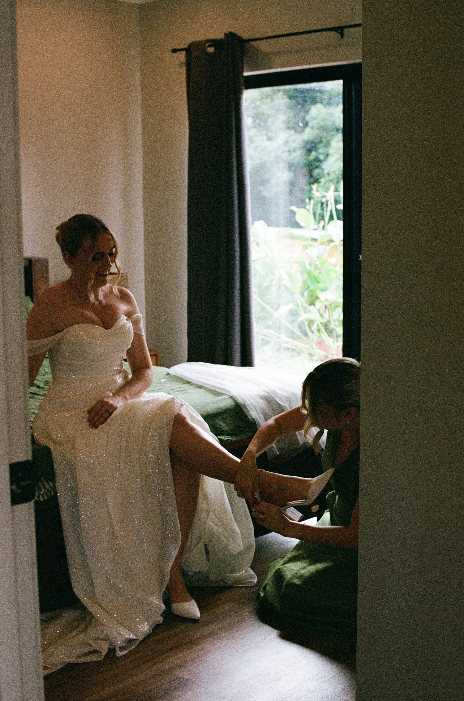 Bridesmaid in green dress kneels to help bride put on white heels before wedding ceremony.