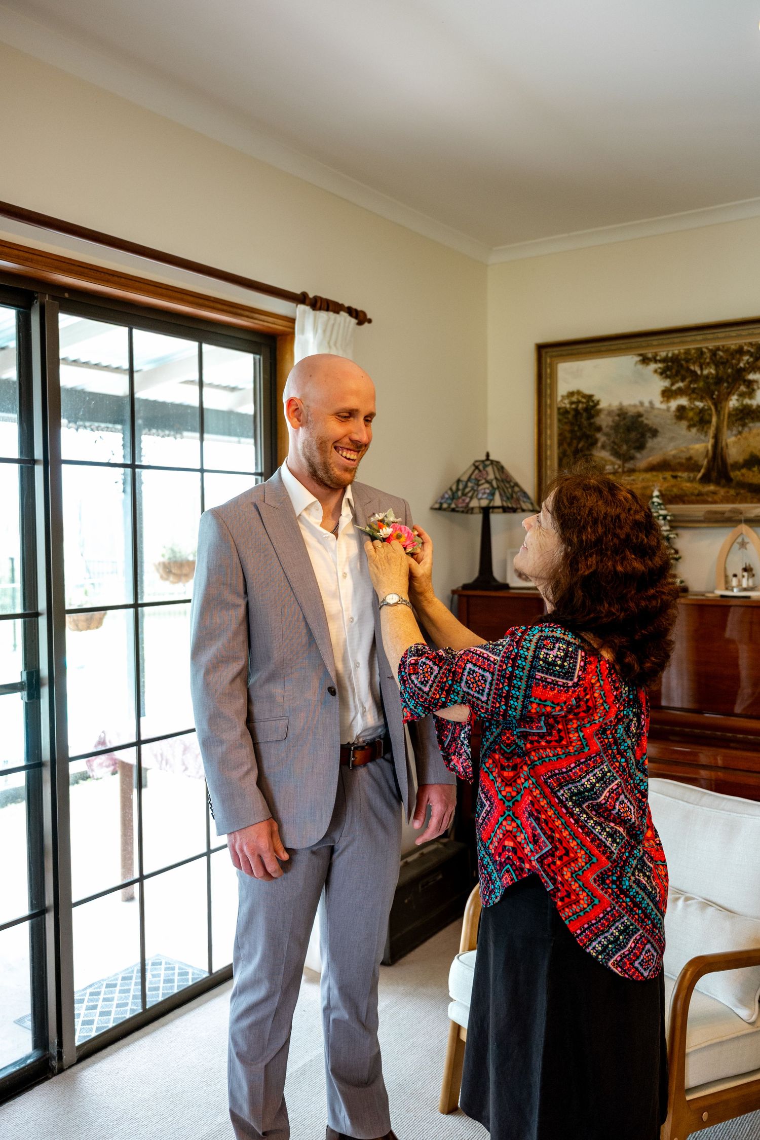 Smiling groom in grey suit receiving boutonniere from woman in colorful patterned jacket.