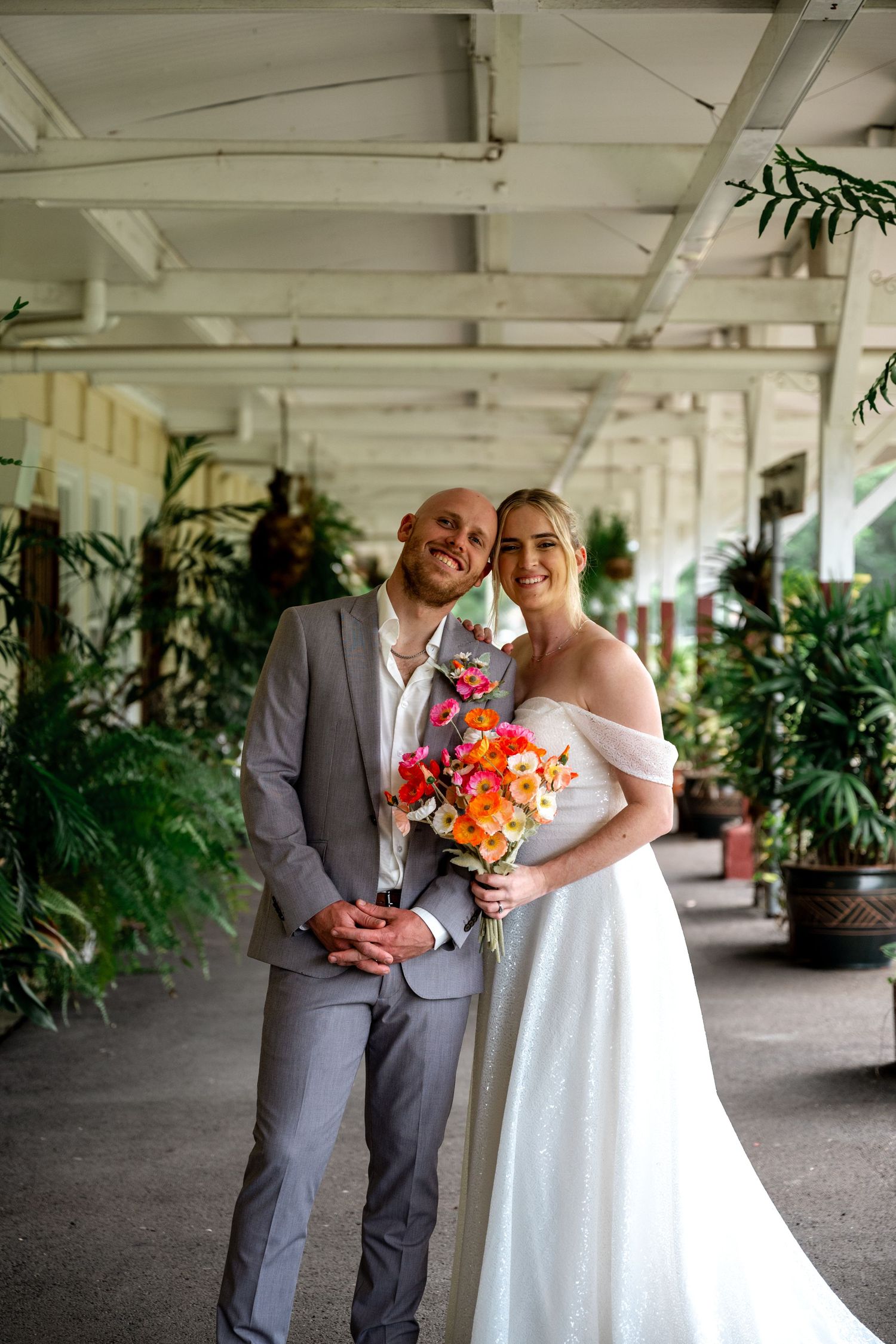 Smiling bride and groom pose together in a covered tropical venue corridor lined with potted plants.