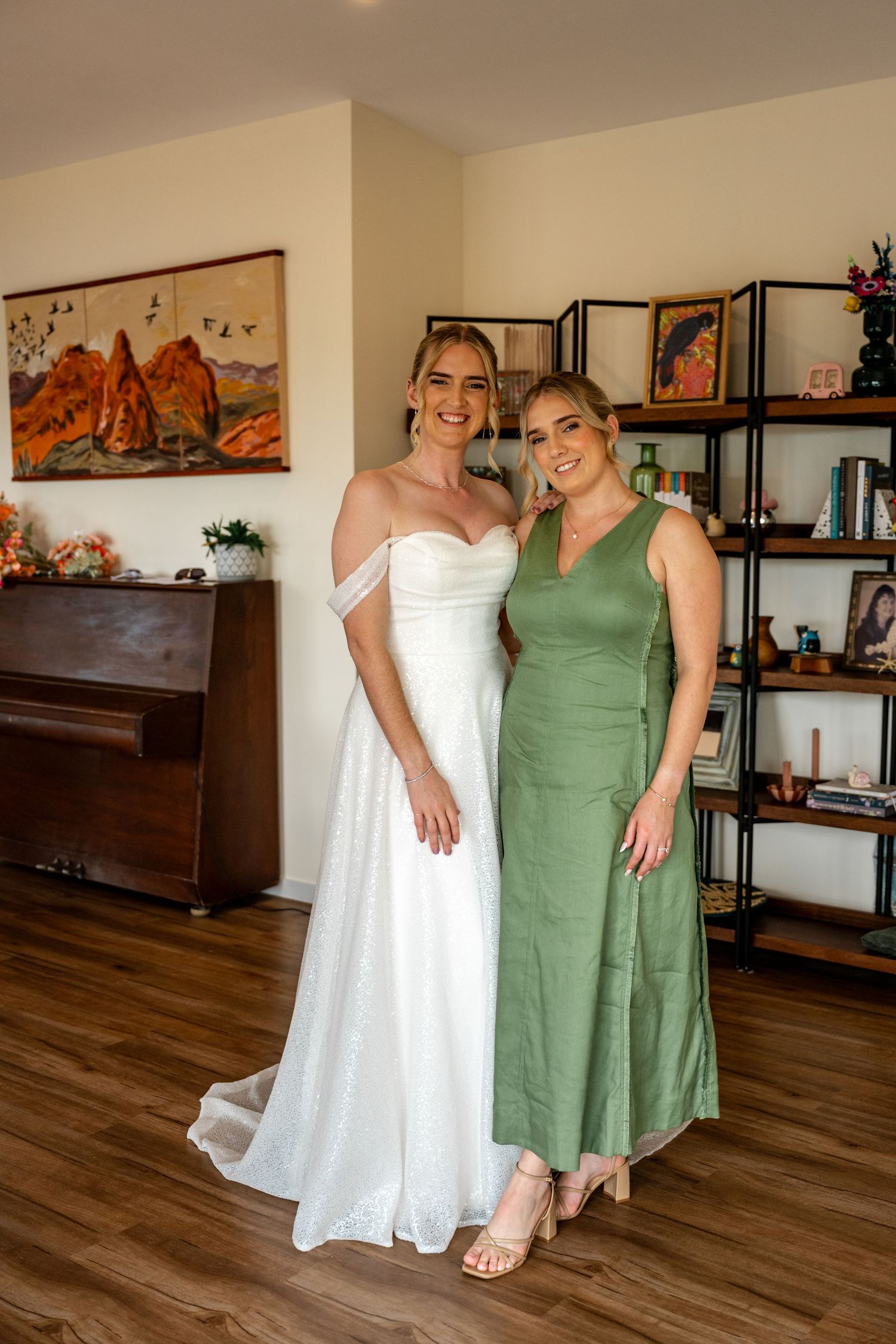 Bride in white off-shoulder gown poses with bridesmaid in sage green dress indoors before wedding ceremony.